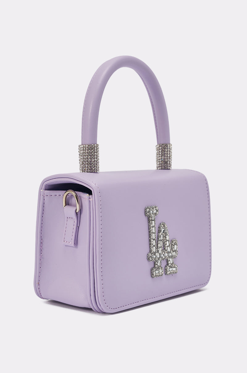 The IN THE CITY APPLIQUE PURSE is a chic lavender mini handbag with a top handle, rhinestone accents on the handle bases, and "LA" in sparkling rhinestones on the front. Perfect to wear as a crossbody bag. 2
