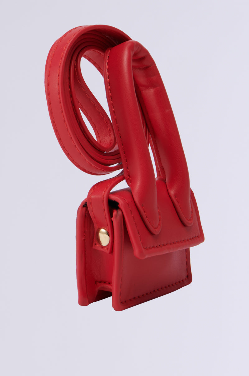 The Runway Chic Bag Bracelet, a compact red faux leather accessory, rests on a smooth surface with its long, coiled strap. The bag is adorned with gold-tone button details on the sides. This multifunctional piece's minimalist design highlights its clean lines and vibrant red hue.