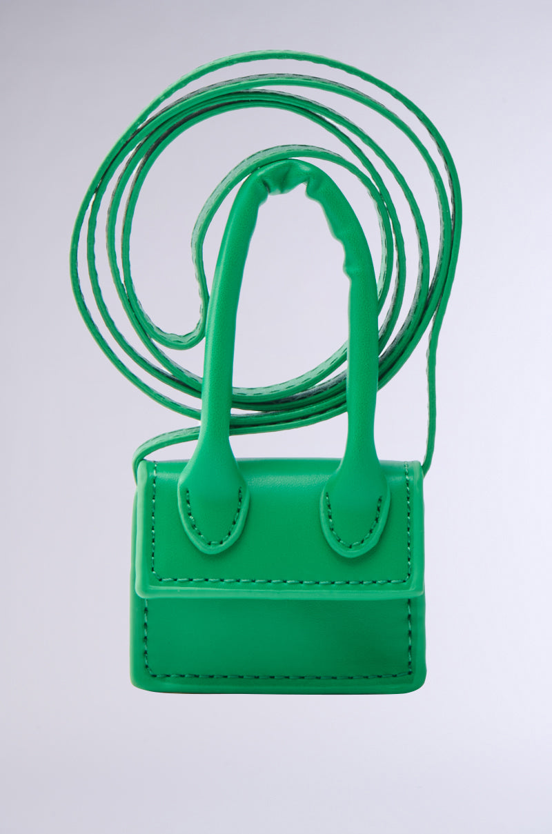 The IN MY BAG BRACELET, a small bright green faux leather handbag with stitched edges, two short top handles, and a long thin strap, is shown against a plain light gray background as a versatile accessory.