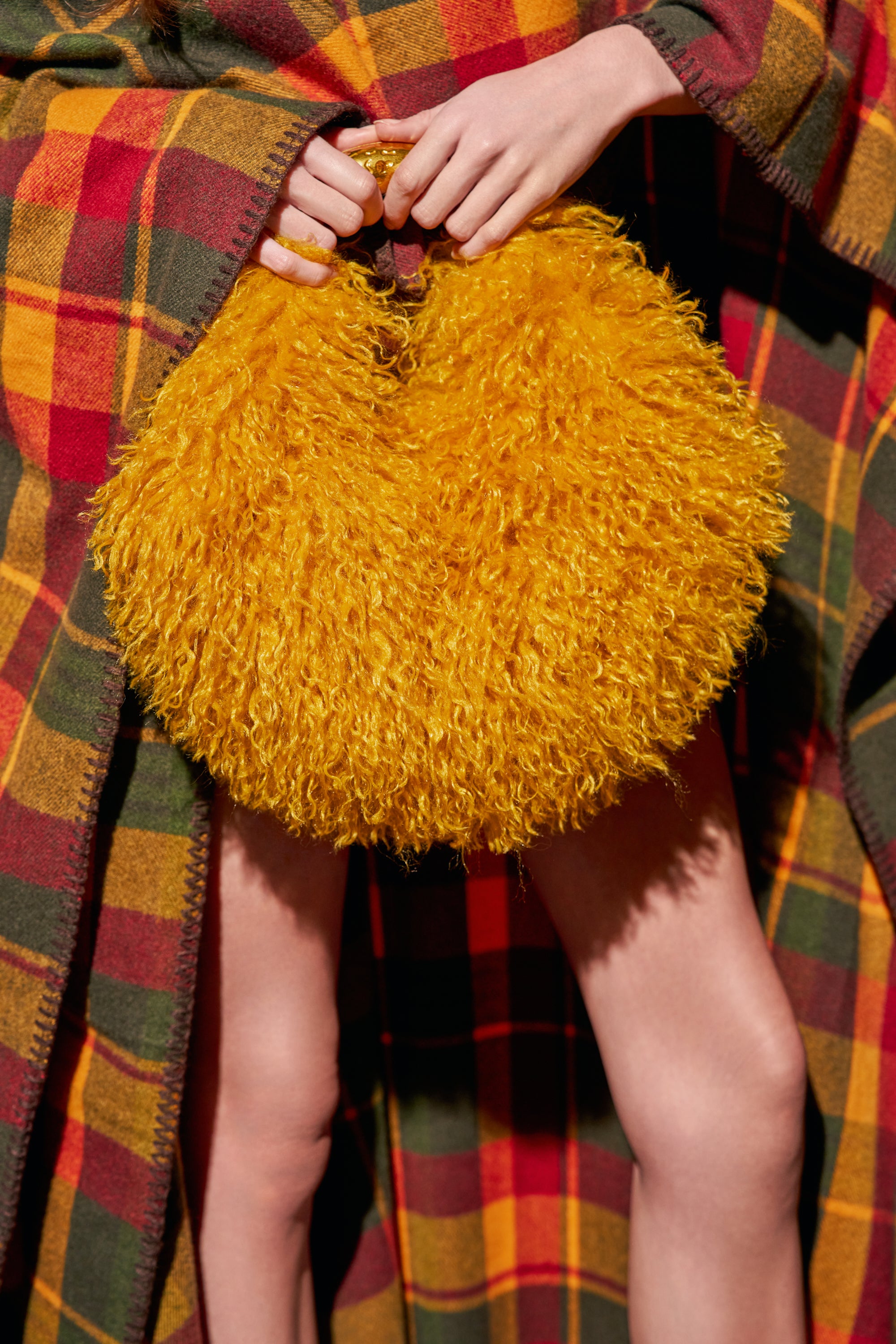 A person holds a round, mustard faux fur SHAGALICIOUS BABY BAG in front of a plaid blanket.