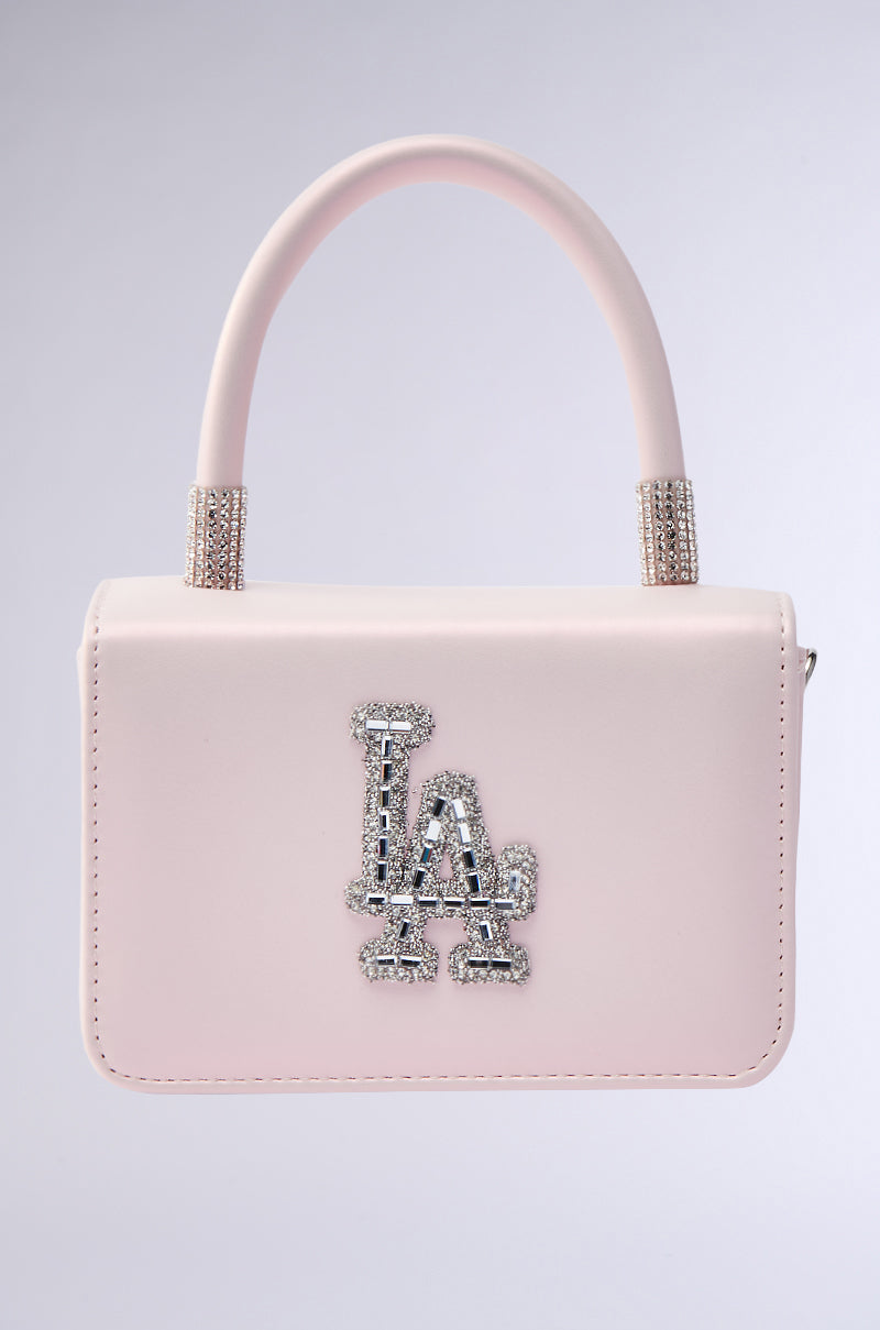 The WHERES MY FLIGHT PURSE is a small, light pink faux leather handbag with a curved handle and sparkling rhinestone accents, featuring "LA" in rhinestones on the front.
