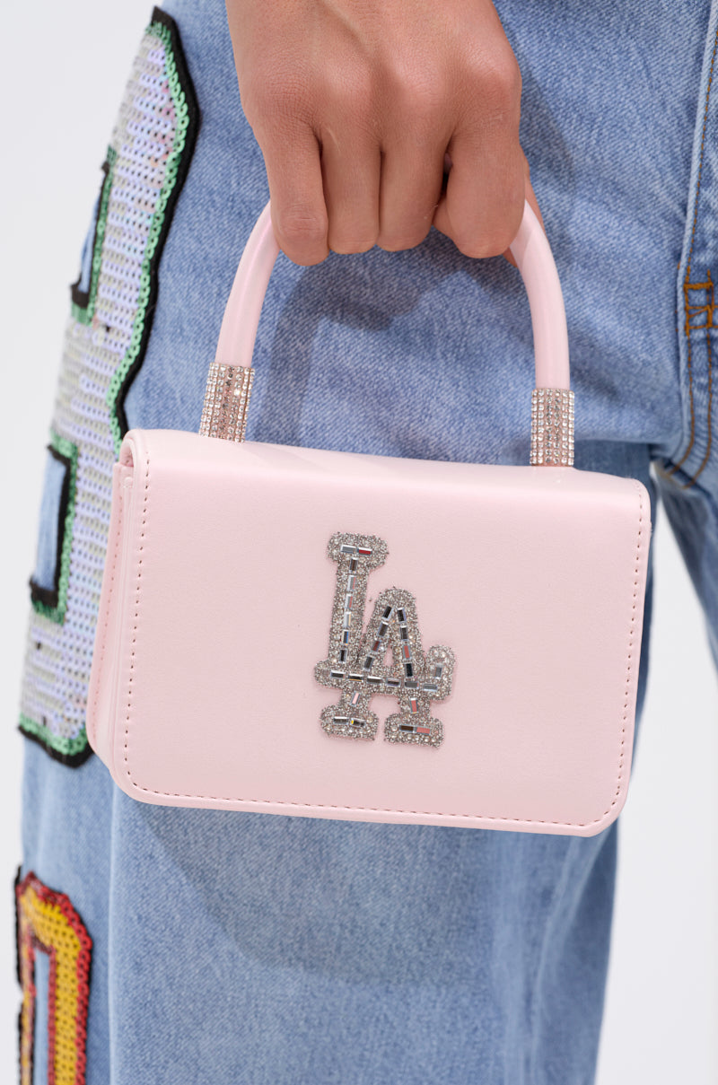 A person holds the WHERES MY FLIGHT PURSE, a small iridescent handbag with an embellished "LA" logo, and wears light blue jeans adorned with colorful sequined patches.