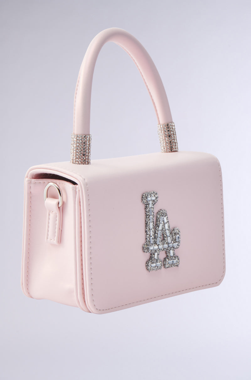 The WHERES MY FLIGHT PURSE is a small light pink faux leather handbag with a top handle, silver rhinestone accents, and a sparkling "LA" logo on the front. Displayed against a plain light gray background.