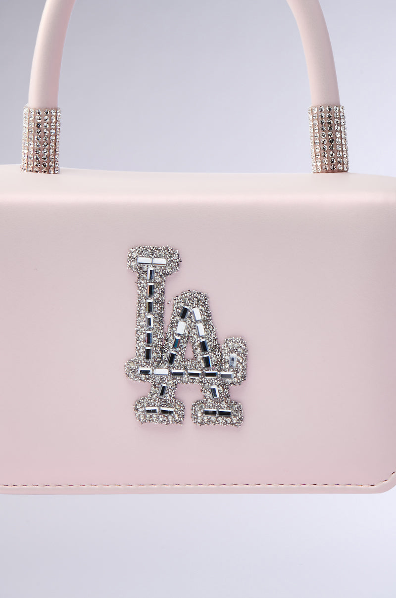 The WHERES MY FLIGHT PURSE is a pale pink faux leather handbag featuring a sparkling, rhinestone-encrusted "LA" logo and rhinestone-accented handles, shown against a light background.