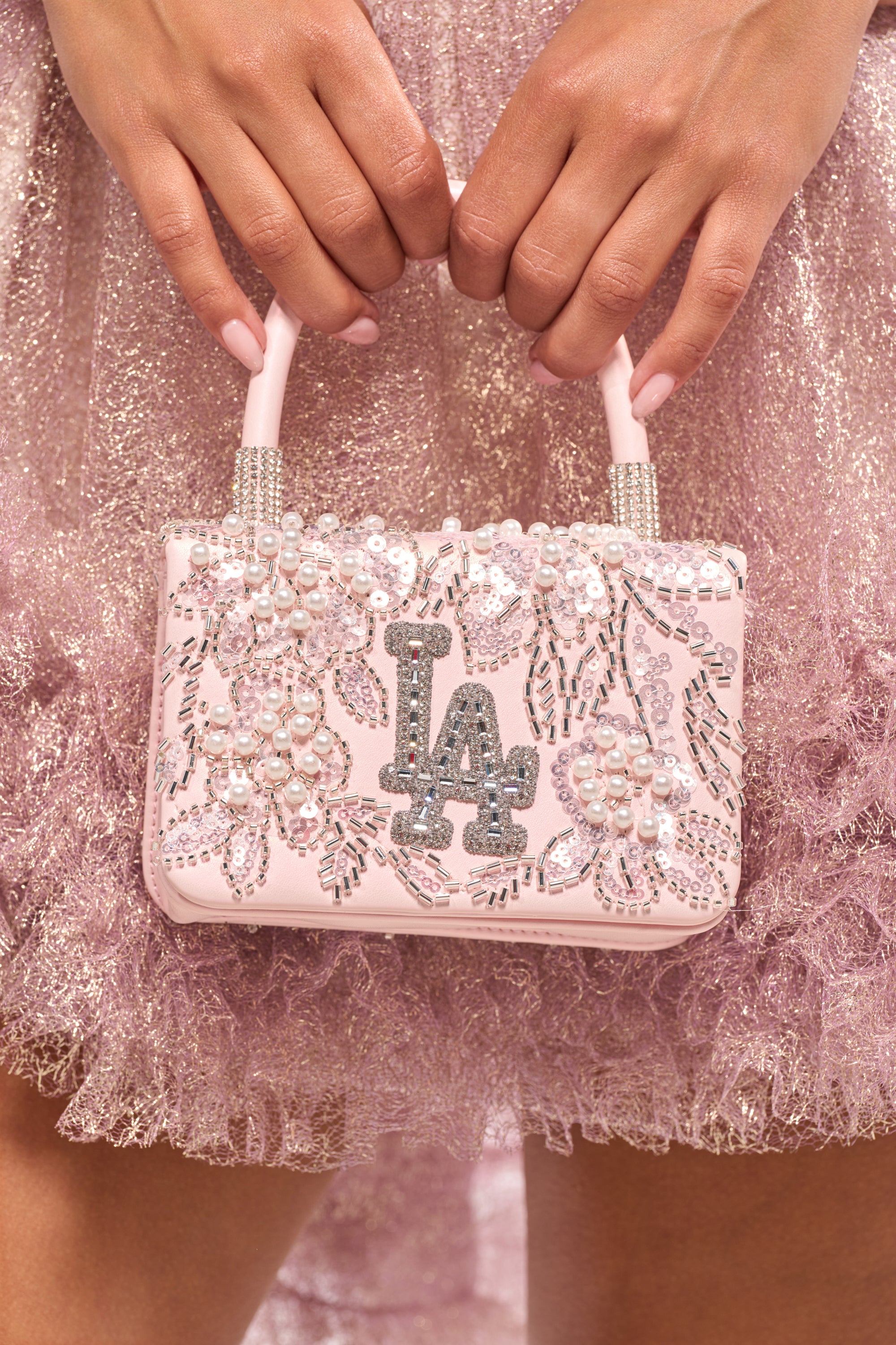 A person in a sparkly pink dress holds the HEARD U PURSE, a rhinestone handbag adorned with pearls, sequins, floral designs, and a glittery "LA" logo. Their nails are painted pale pink to match. 1