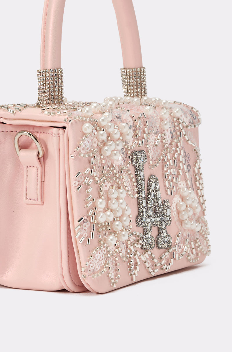 The HEARD U PURSE is a small, pale pink faux leather mini handbag with pearl and crystal bead embroidery, a bold silver "LA" logo on the front, and crystal-embellished handles near the base. 4