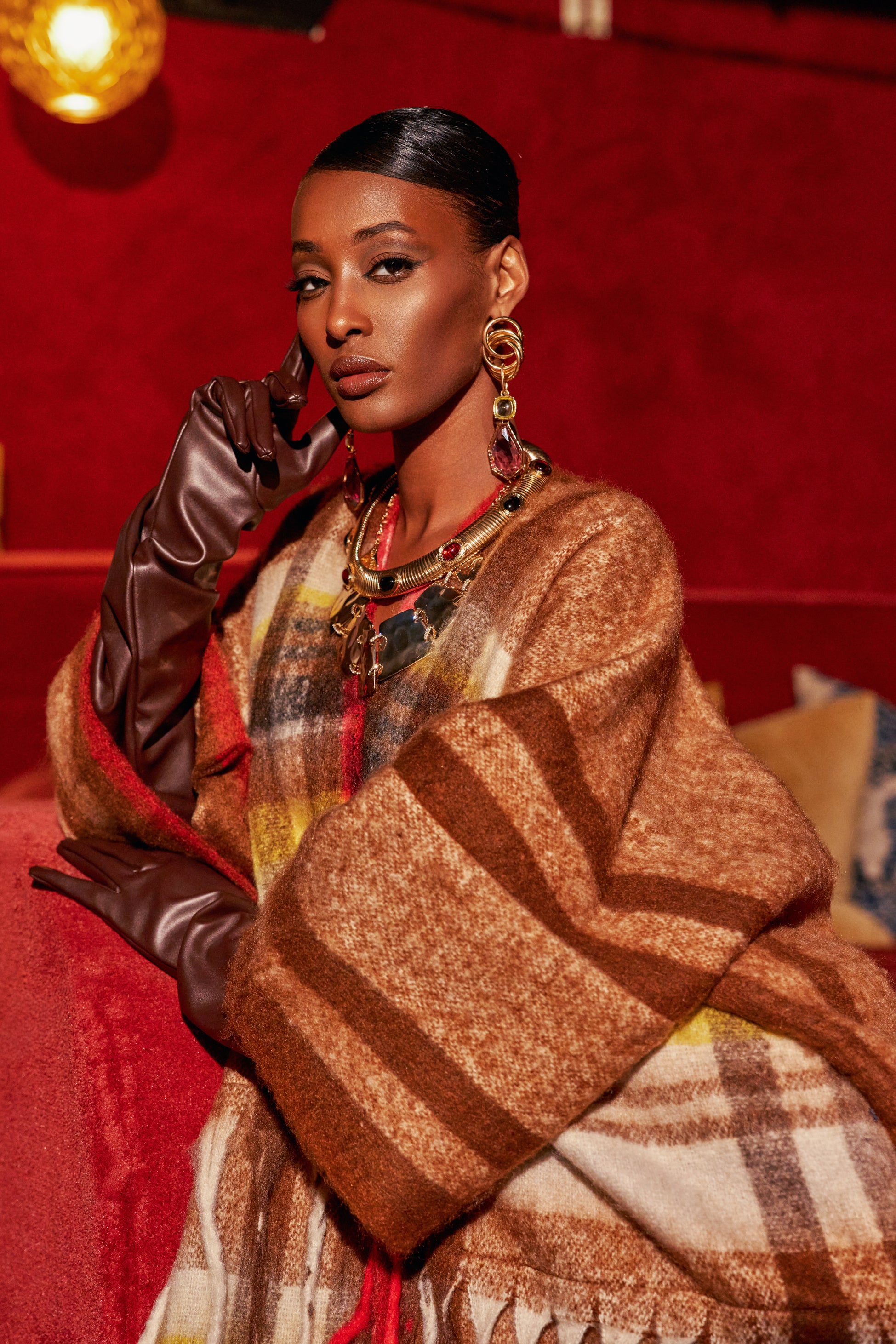 A person wearing THAT GIRL LONG GLOVES in chocolate brown faux leather, paired with a plaid shawl and large gold statement jewelry.