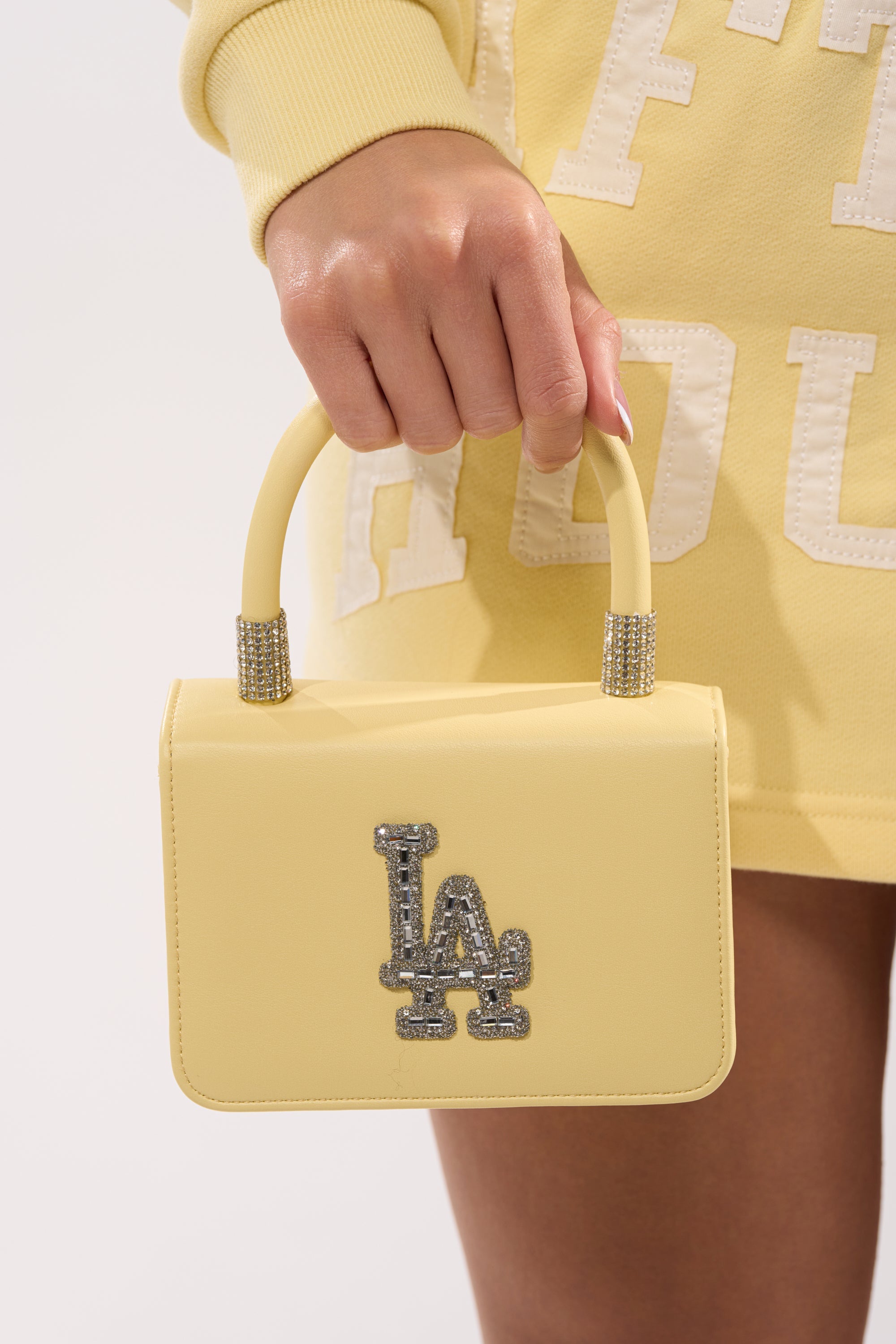 A person in a yellow outfit holds the IN THE SUN BLING PURSE—a mini yellow top-handle evening bag featuring a rhinestone-encrusted "LA" logo and decorative accents near the base of the handle. 2
