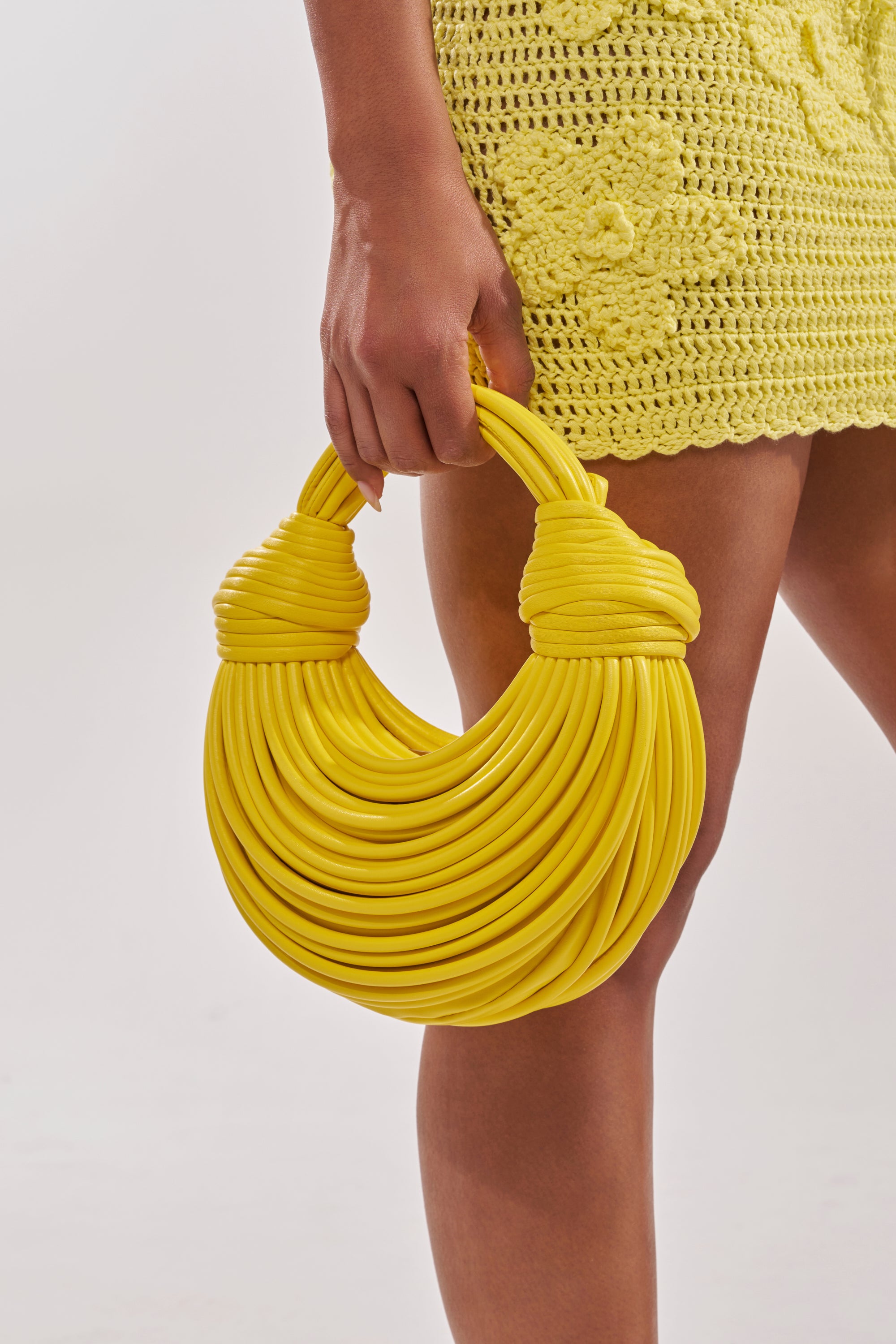 A person holds the BANANA SPLIT WHO PURSE, a bright yellow faux leather handbag with wrapped handles—an ideal summer accessory—while wearing a yellow crocheted skirt against a white background. 1