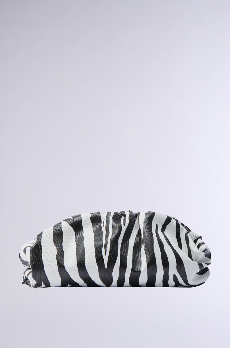The INTO THE WILD ZEBRA CLUTCH, featuring a black and white zebra-striped pattern and crafted from faux leather, is displayed against a plain gray background. Its soft, folded design gives it the appearance of a pillow or pouch.