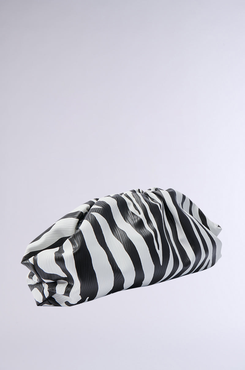 The INTO THE WILD ZEBRA CLUTCH, crafted from faux leather with an eye-catching black and white zebra pattern, is folded and resting on a light gray background. Its unique design showcases rippled horizontal stripes, giving it a textured appearance.