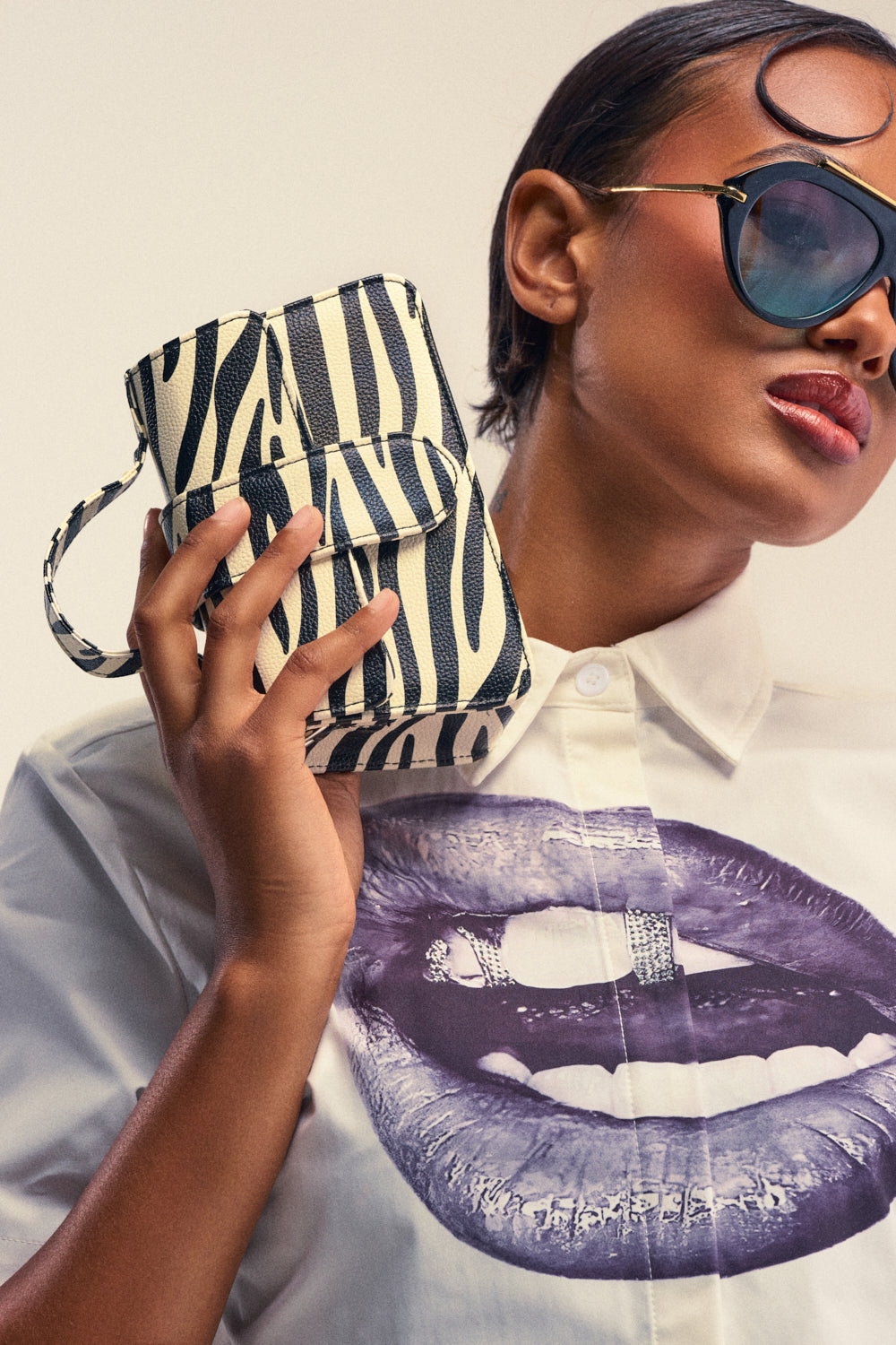 A person wearing sunglasses and a shirt with a graphic of lips holds the LETS GET WILD ZEBRA BAG—a structured mini bag featuring a zebra print and a removable chain strap—next to their face against a plain background.