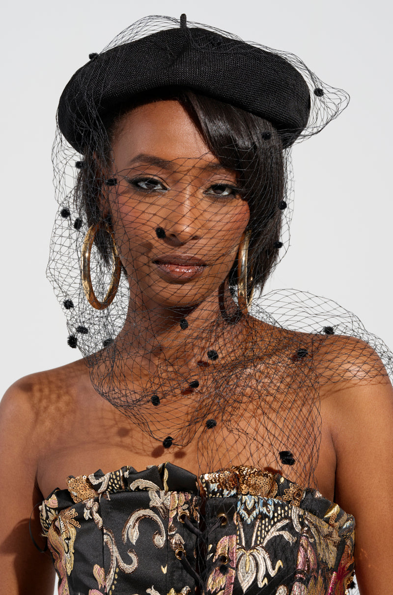 A woman confidently poses, her silhouette adorned with the PUT ME ON THE COVER EDITORIAL HAT, featuring a black beret accented by a mesh net veil and playful pom-poms. Large hoop earrings complement the colorful, ornate pattern of her strapless dress. Her poised expression is highlighted against a plain backdrop. 0