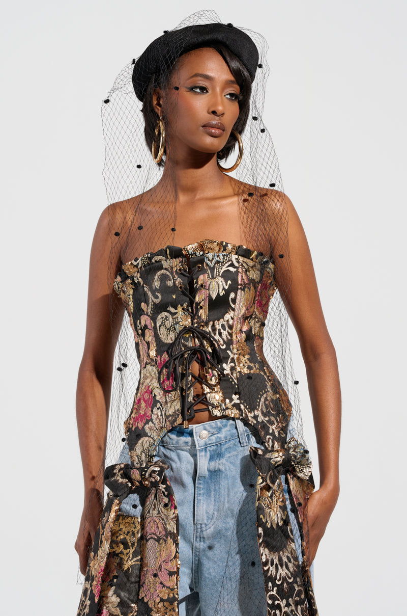 A woman models a chic ensemble, featuring a strapless lace-up floral corset paired with high-waisted blue jeans. She accessorizes with large hoop earrings and the PUT ME ON THE COVER EDITORIAL HAT, elegantly adorned with a black mesh net veil. Her pose exudes confidence against a neutral backdrop. 1