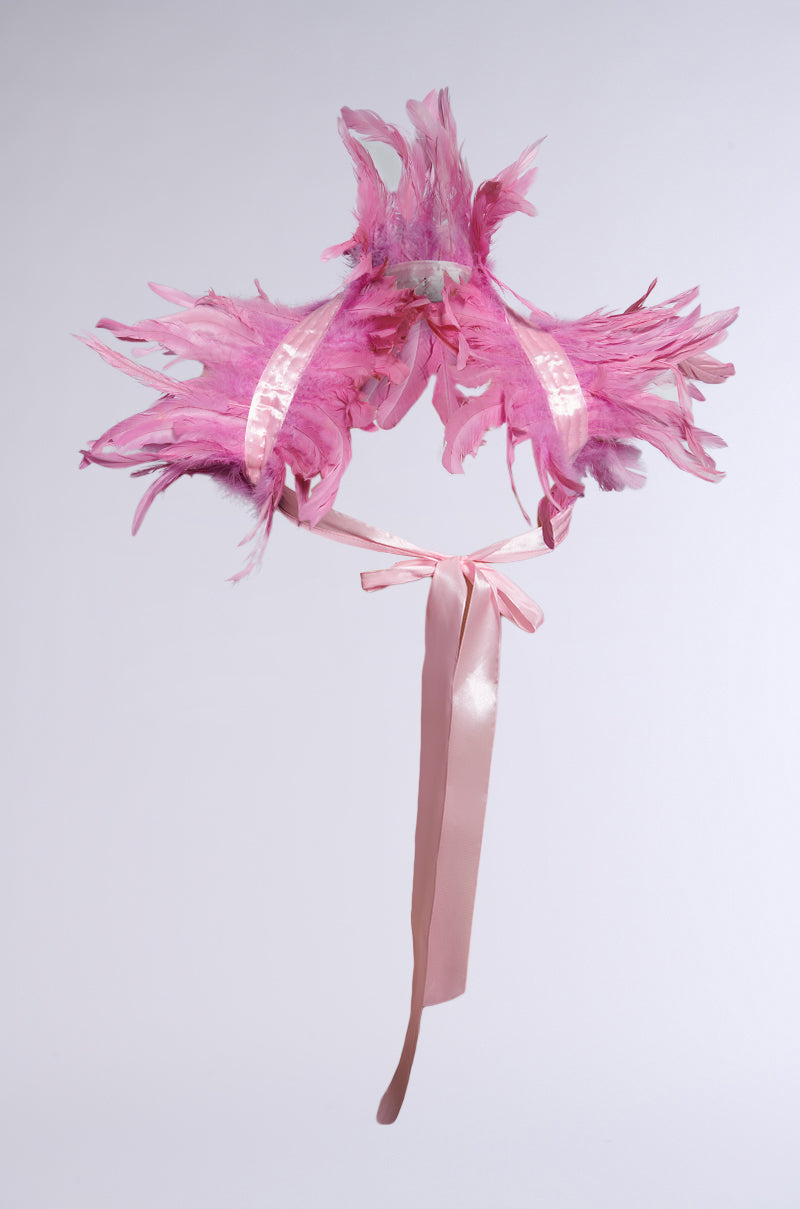 The HERE FOR THE DRAMA FEATHER BOLERO is a decorative headpiece featuring vibrant pink feathers and pink ribbon ties against a plain light gray background. 3
