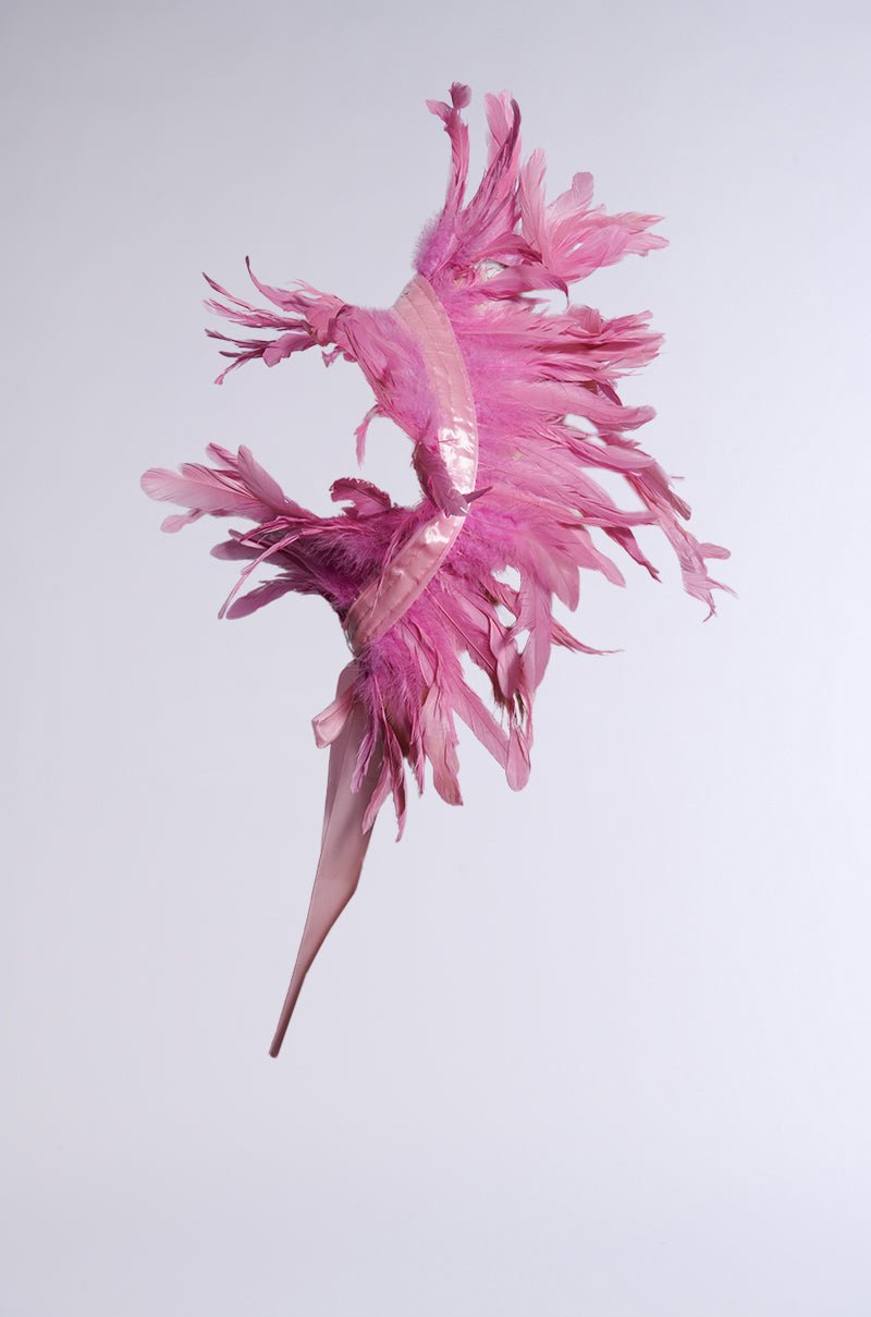 The HERE FOR THE DRAMA FEATHER BOLERO is a striking pink headpiece with dramatic elongated feathers, an intricate design, and a glossy ribbon-like band at the base, creating a bold statement on any plain background. 4