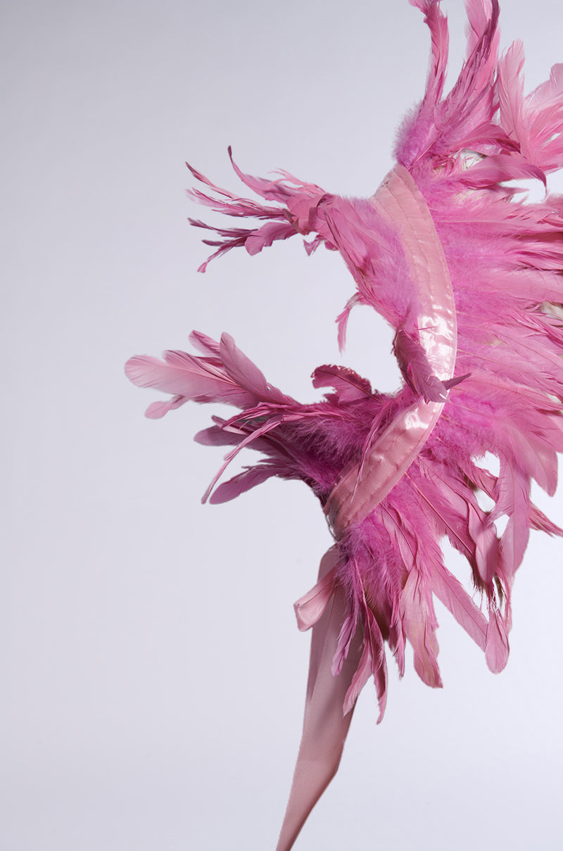 The HERE FOR THE DRAMA FEATHER BOLERO is a striking pink feathered headpiece with intricate layering and a reflective ribbon accent, set against a light gray background. Its feathers fan out dynamically, exuding movement and elegance. 5