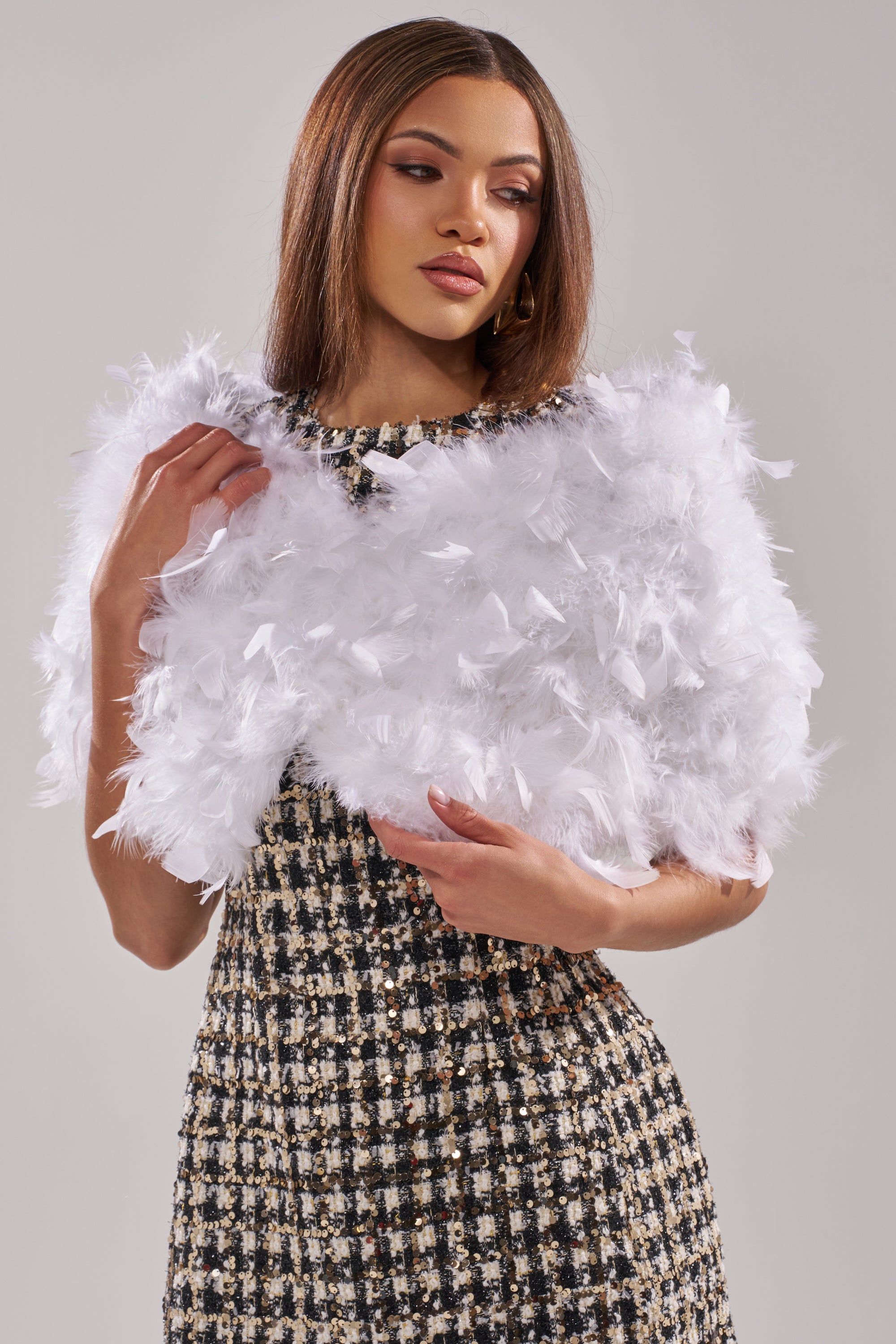 A woman stands against a plain light gray background, draping the TRUDY FEATHER WRAP—a striking white feather statement piece—over a black and white tweed dress.