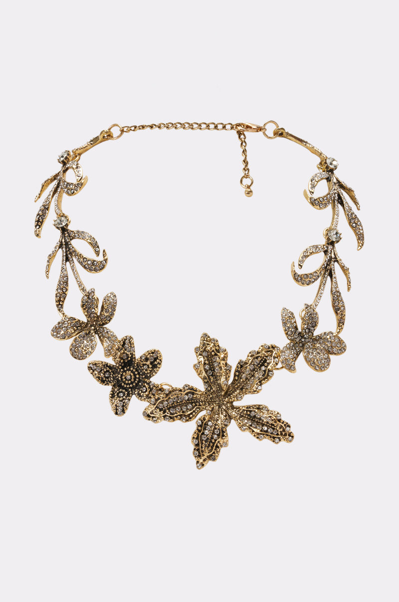 The CRYSTAL PETAL STATEMENT NECKLACE showcases intricate floral and leaf motifs adorned with sparkling decorative stones, reminiscent of a rhinestone statement piece, and features an adjustable chain clasp. Presented against a plain white background.