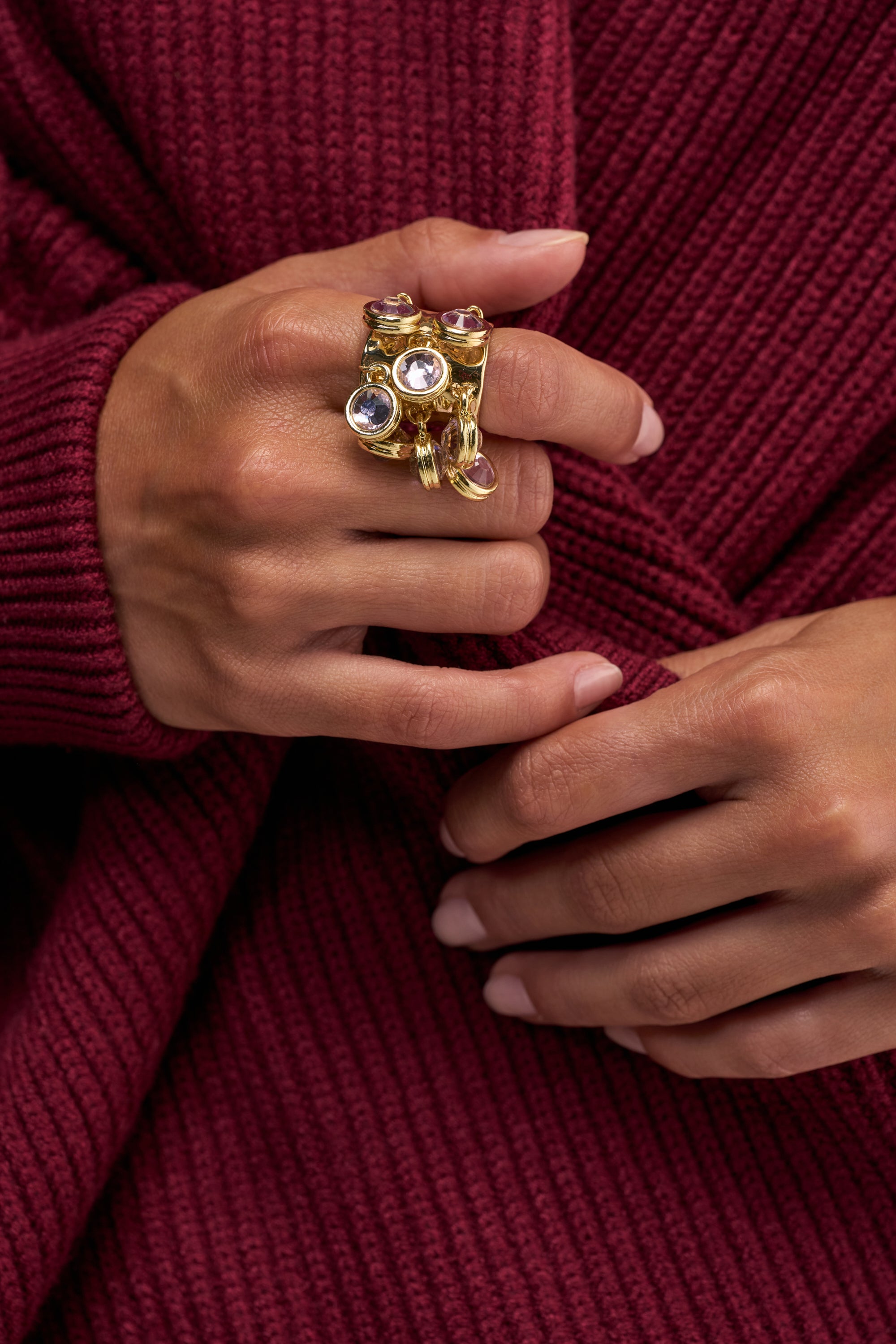 Wearing a textured maroon sweater, an individual showcases several gold rings adorned with vibrant gemstones on their hand, including the GILDED GEM CLUSTER RING—an adjustable gold piece that serves as a striking statement accessory—as their fingers gently touch their opposite wrist.