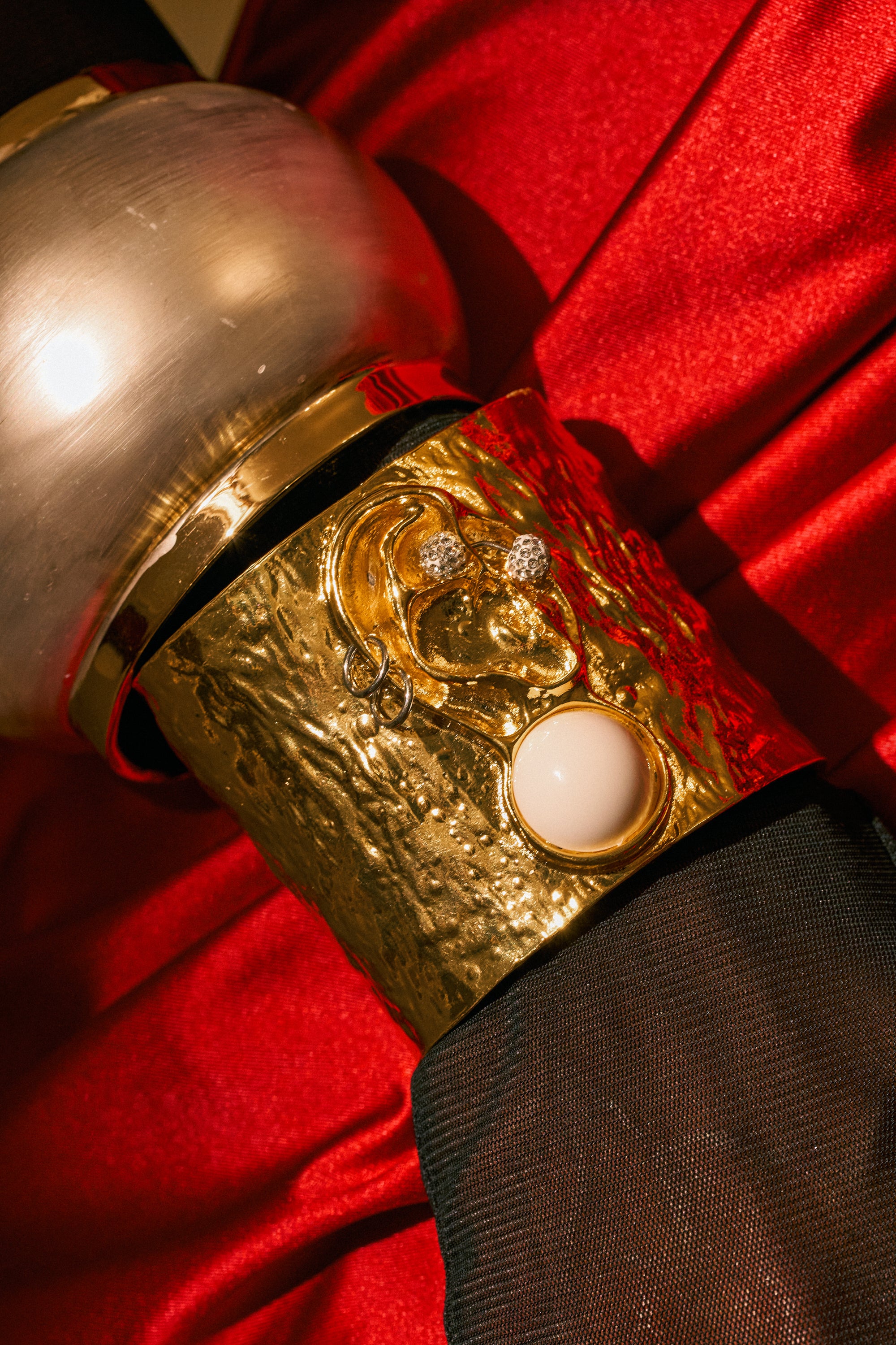 A close-up of the LISTEN CLOSELY METAL CUFF IN GOLD, featuring a face design with diamond-like accents and a pink gemstone, worn over a sheer black sleeve and red satin fabric. The hammered gold surface adds texture and depth, beautifully catching the light.