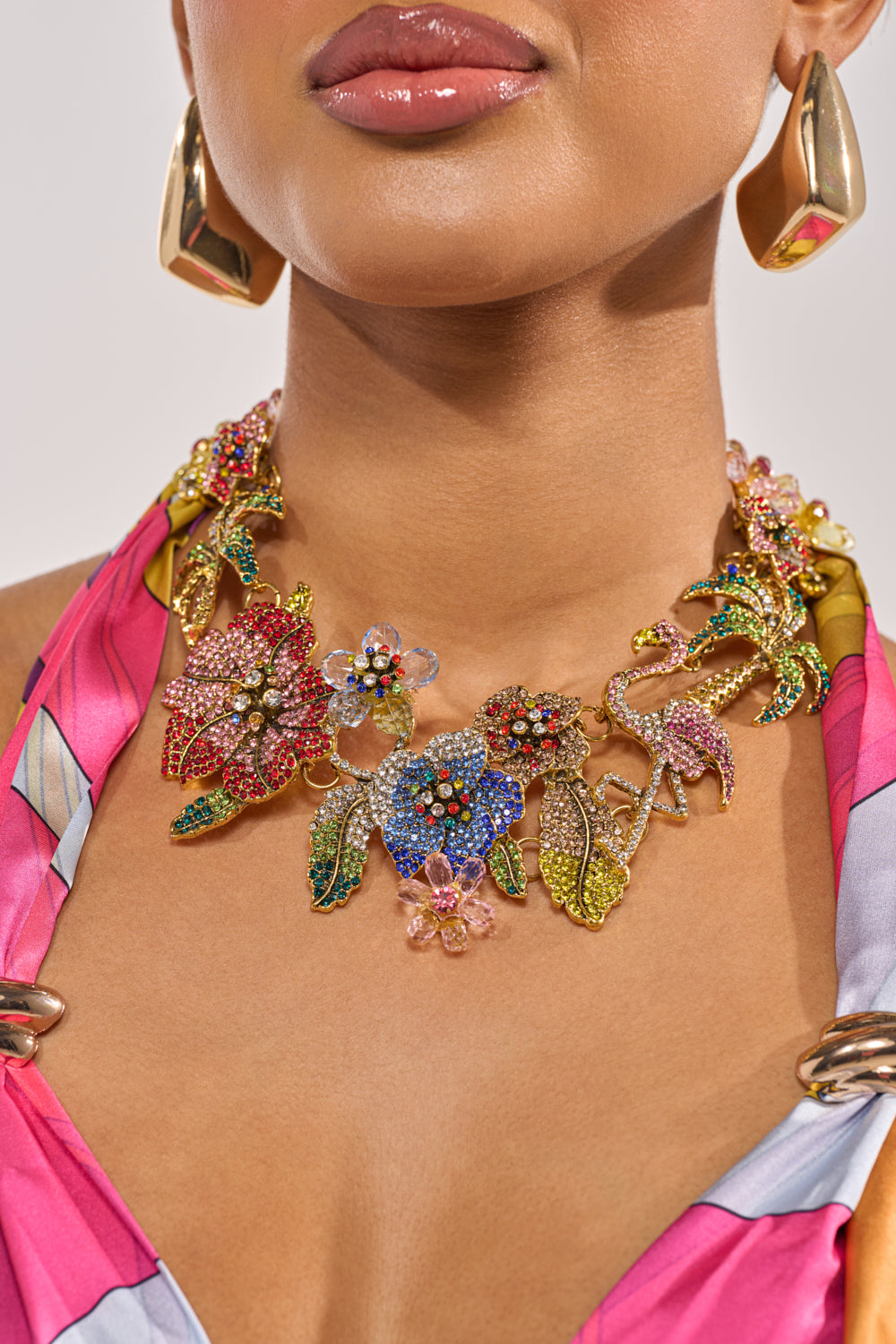 A woman wears the IN THE GARDEN STATEMENT NECKLACE with vibrant rhinestones, floral and bird designs, plus colorful gems. She pairs it with large gold earrings and a pink patterned top; her glossy lips complete the resort-ready look. 1