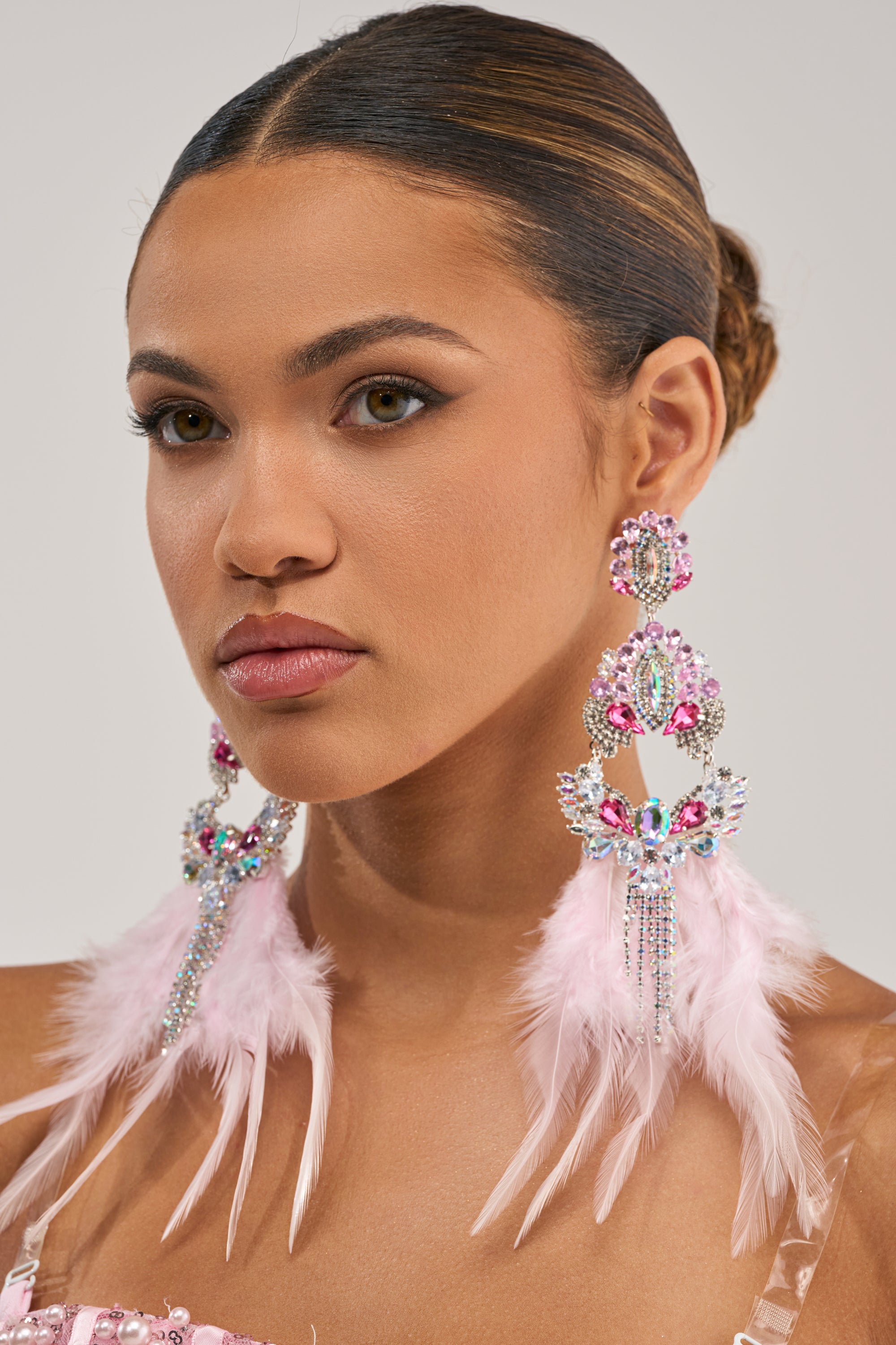 A woman with sleek dark hair in a bun wears the CARNIVAL EARRING, featuring pink rhinestones, crystals, and long feather drops. She looks calm and elegant.