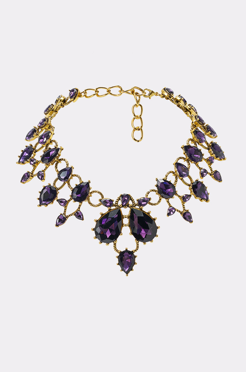 The ON TREND STATEMENT NECKLACE is a gold evening piece with large and small faceted purple gemstones in a symmetrical ornate design, featuring an adjustable chain clasp—ideal for formal events.