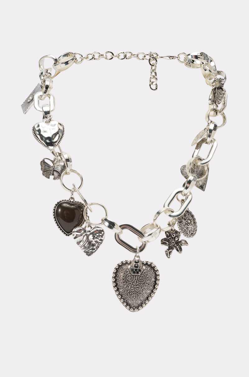 The SUCH A CHARMER CHOKER NECKLACE features a bold silver chain with large links, adorned with an array of ornate, engraved heart-shaped and floral charms. Displayed on a plain white background, this statement necklace is perfect for elevating any look.