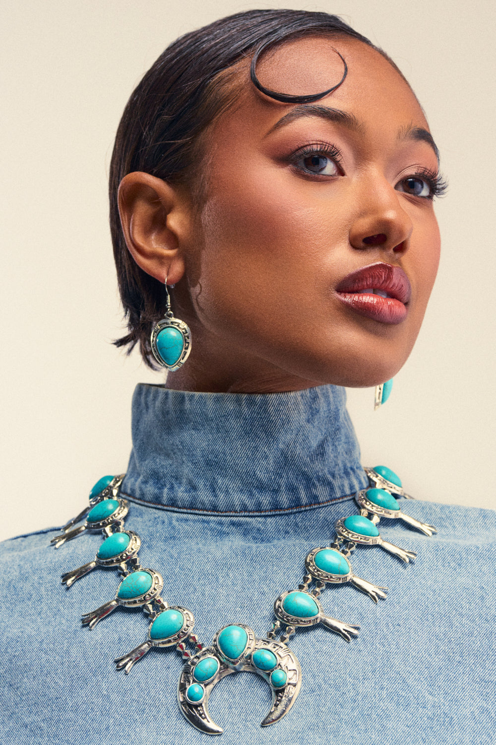 Wearing the MALINA EMBELLISHED TURQUOISE NECKLACE SET, which features a southwestern statement necklace adorned with crescent and bird motifs and turquoise stones, along with matching festival-inspired turquoise and silver earrings, a woman in a high-collared denim top gazes confidently to the side.