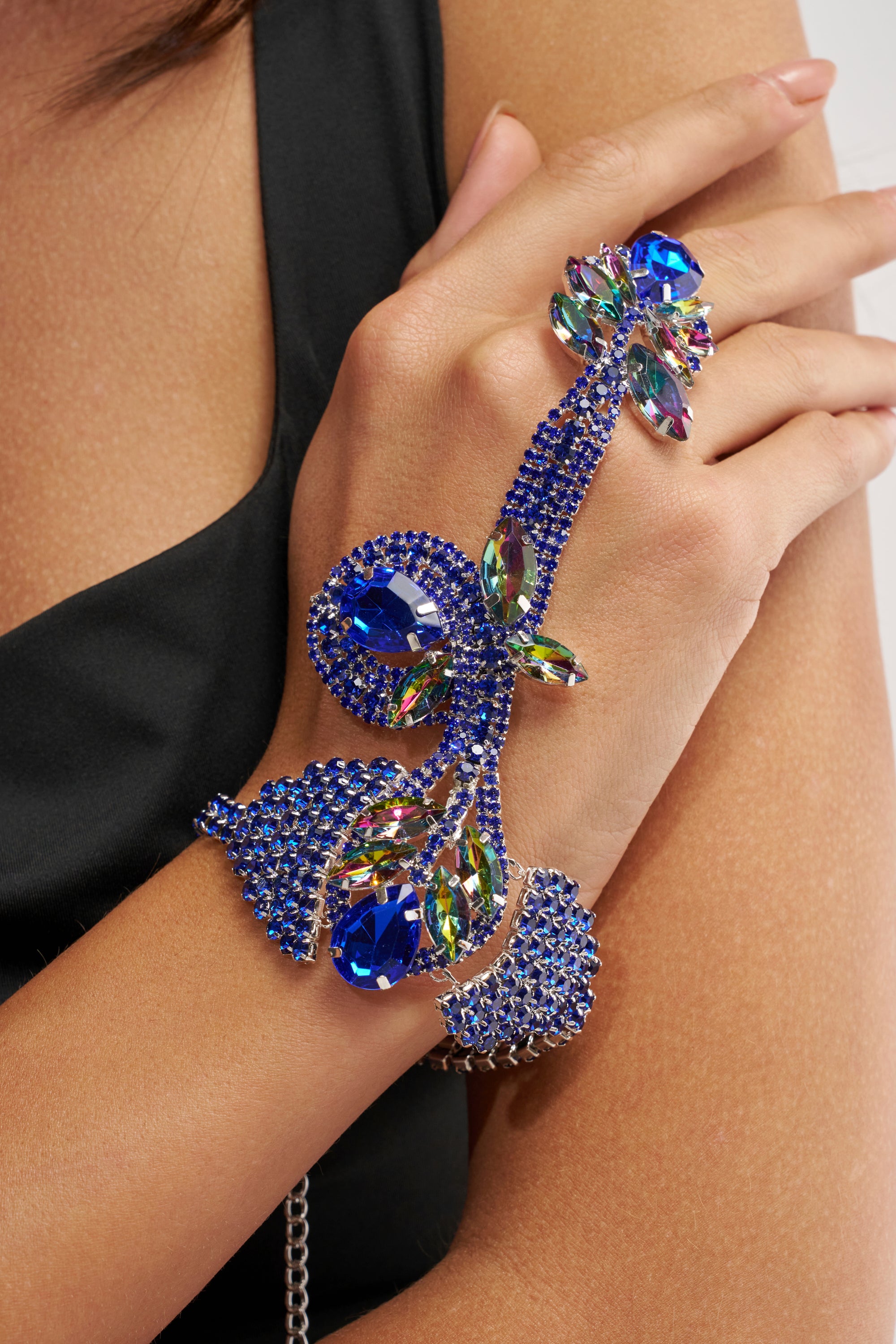 A person wears the WATCH ME BRACELET, an ornate blue and multicolored jeweled handpiece shaped like a vine, with stones that adorn the wrist and extend across the hand—a striking accessory paired with sleek black attire.
