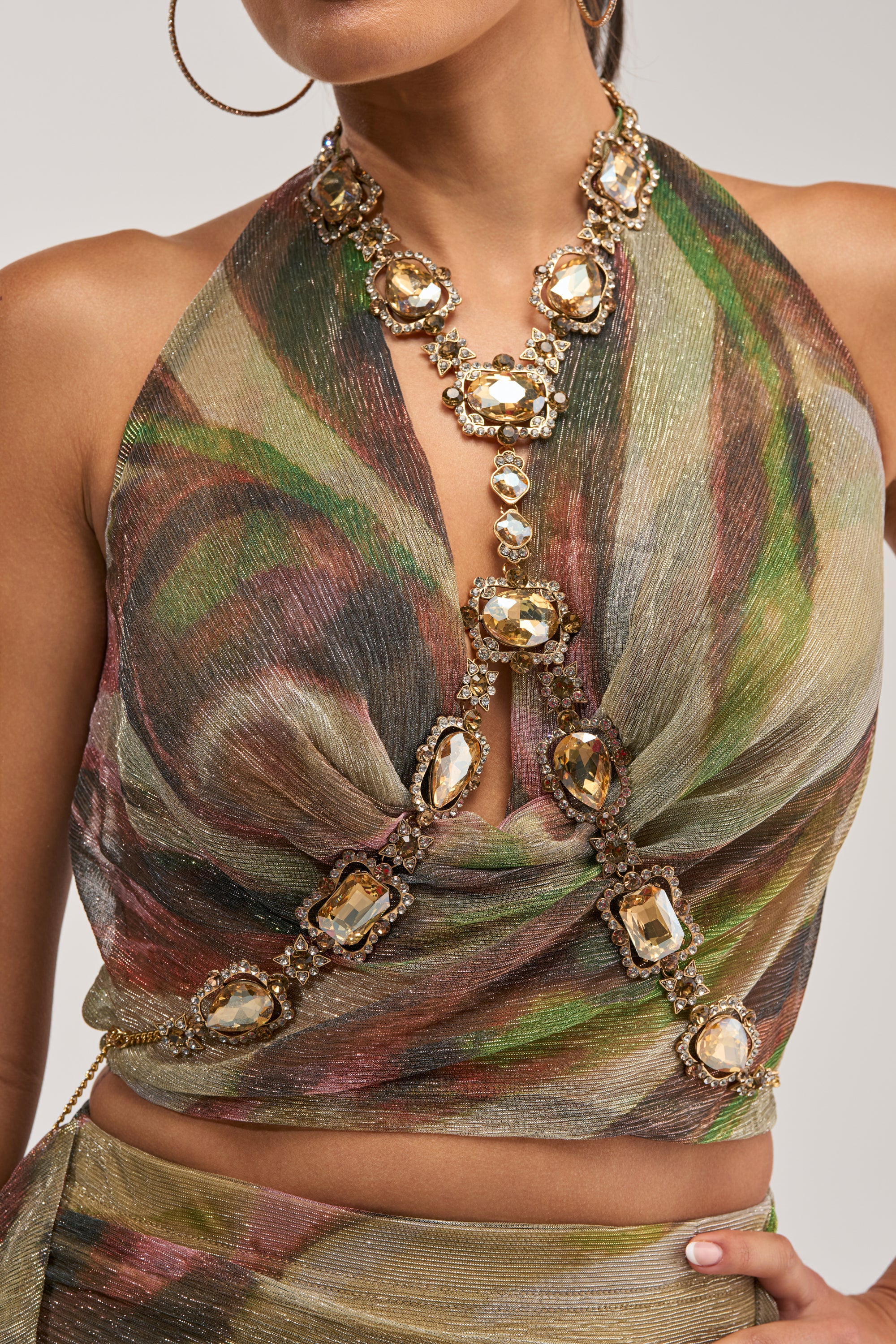 A woman is wearing a deep V-neck halter crop top in multicolor swirl metallic fabric, accessorized with a gold statement gem necklace showcasing large champagne crystals cascading down the front, and styled with the GOING OUT BODY CHAIN for the ultimate touch of festival jewelry.