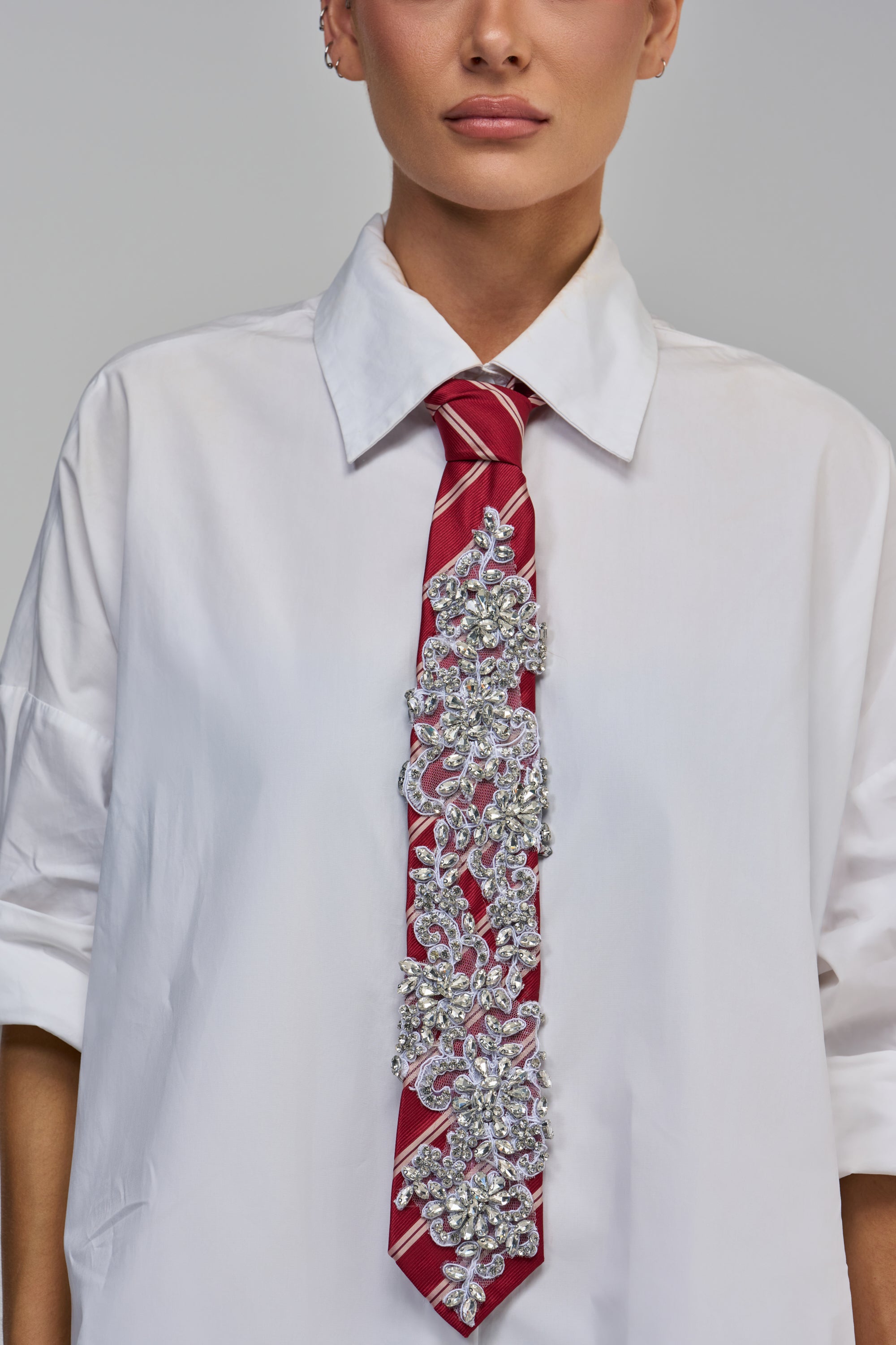 A person wears a SPARKLE BUSINESS EMBELLISHED TIE, a burgundy striped tie featuring a large, intricate floral design made of clear crystals, over a white button-down shirt.