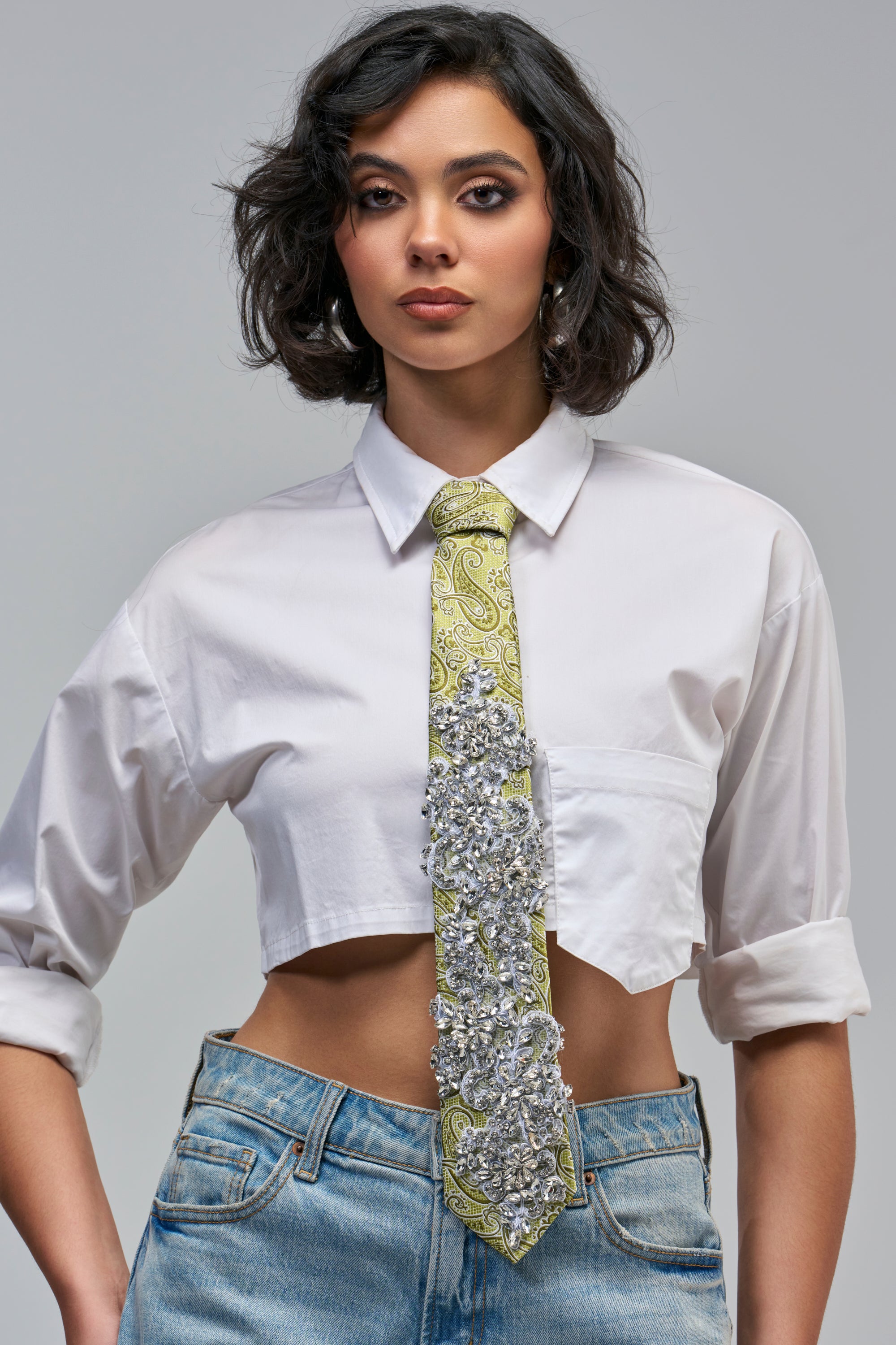A person wears a white cropped collared shirt and blue jeans with a TIE ME UP EMBELLISHED PAISLEY NECKTIE in lime green, heavily embellished with silver crystals and a silver brooch, for a statement officewear look.