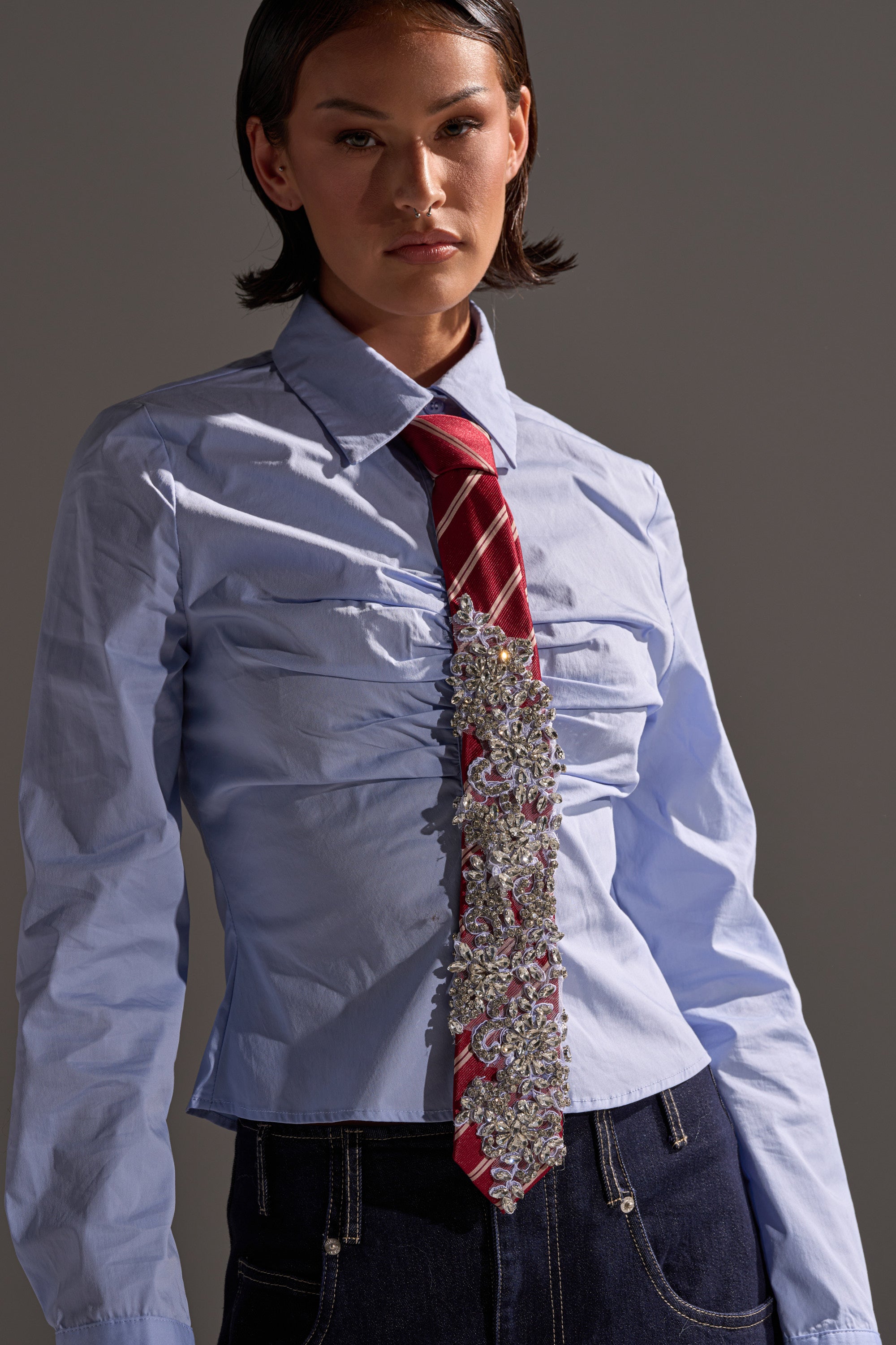 A person wearing a light blue ruched button-up shirt, dark denim jeans, and the NOT YOUR TYPICAL TIE IN RED stands against a neutral background. 0