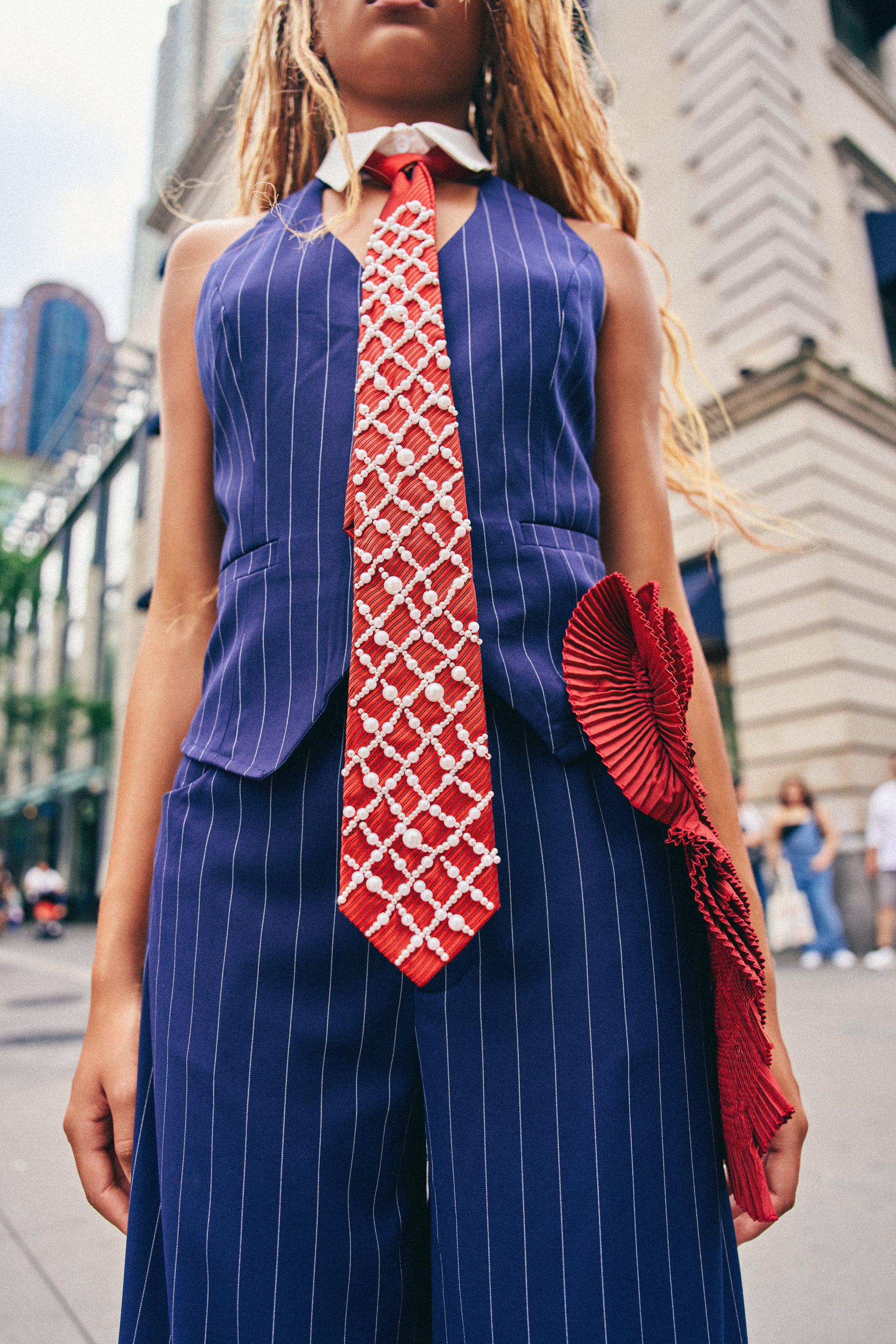 A person in a blue pinstripe suit, wearing the BUSINESS BADDIE TIE—a bold red and black striped satin tie—paired with a large red ruffle accessory, stands outdoors near a city building.