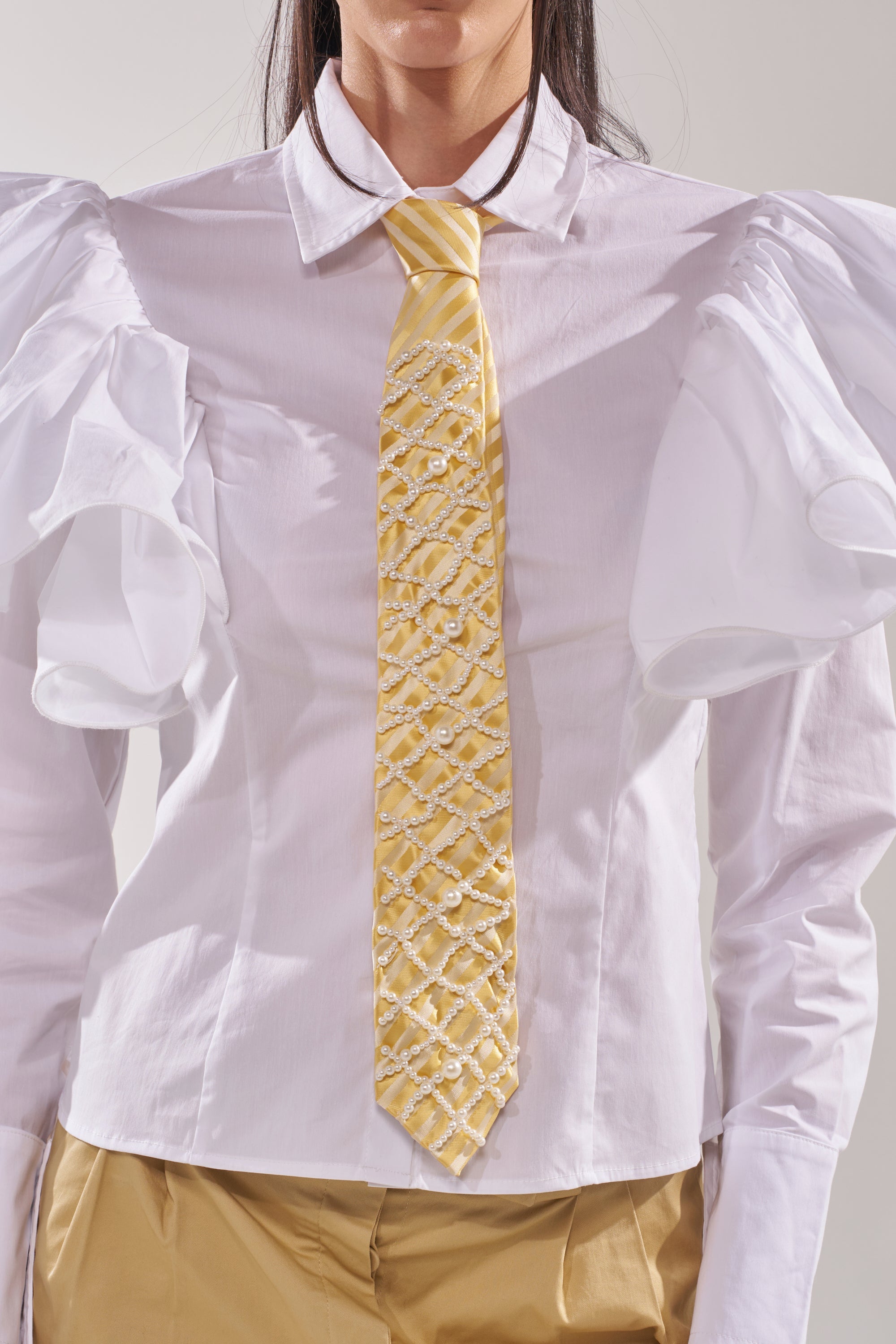 A person wears a white shirt with large ruffled sleeves, light brown pants, and the FEMME APPLIQUE TIE—a butter yellow necktie with a crisscross beaded design. The image is cropped at the shoulders and waist. 2