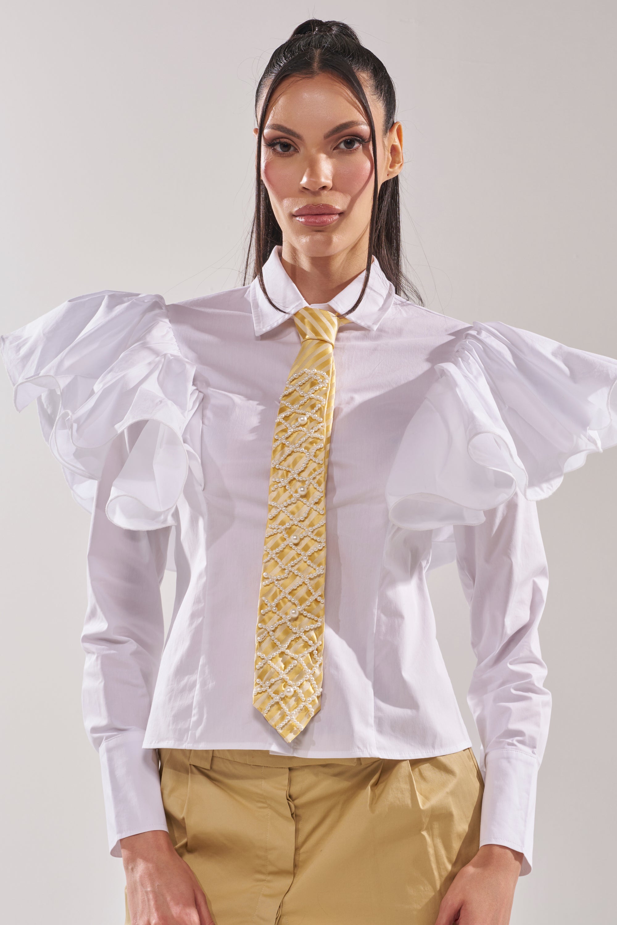 A woman faces the camera against a plain background, wearing the FEMME APPLIQUE TIE with a white blouse featuring dramatic ruffled sleeves and gold pants. 7