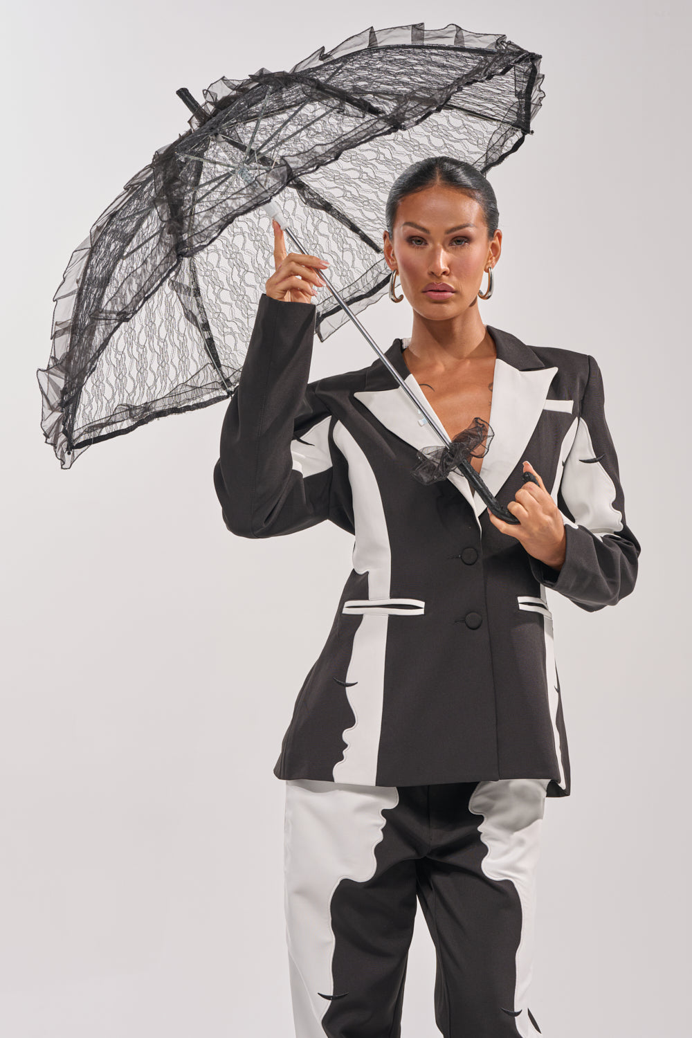 A woman in a black and white suit holds the UNDER MY SPELL LACE UMBRELLA with a scalloped edge. Her hair is pulled back, she wears hoop earrings, and she poses confidently against a plain light background. 1