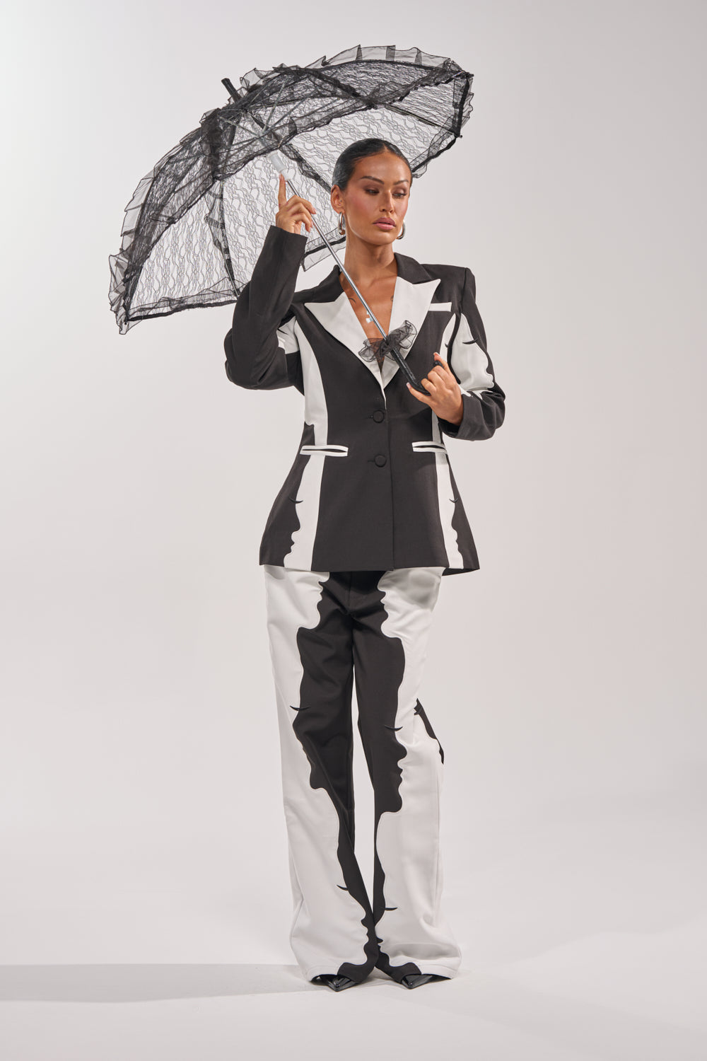 A person holds the UNDER MY SPELL LACE UMBRELLA with a black lace canopy, dressed in a bold black-and-white jagged-pattern suit with wide-leg pants, set against a plain white background. 3