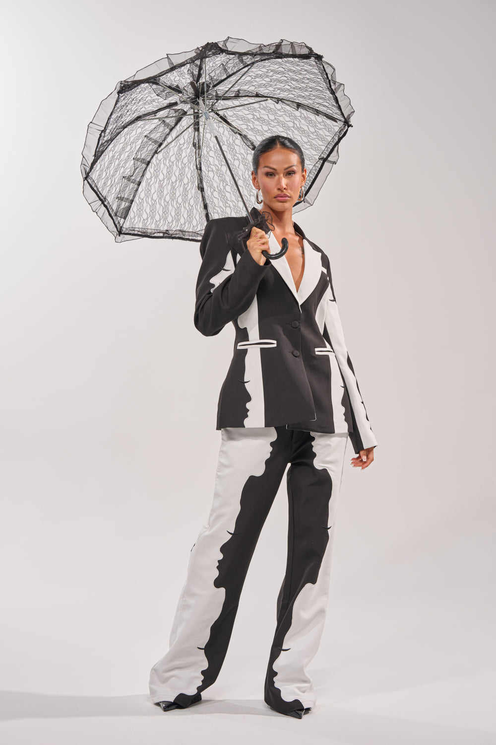 A woman stands confidently against a plain background, holding the UNDER MY SPELL LACE UMBRELLA with its black floral lace canopy, and wearing a bold black-and-white suit featuring an abstract mirrored pattern. 4