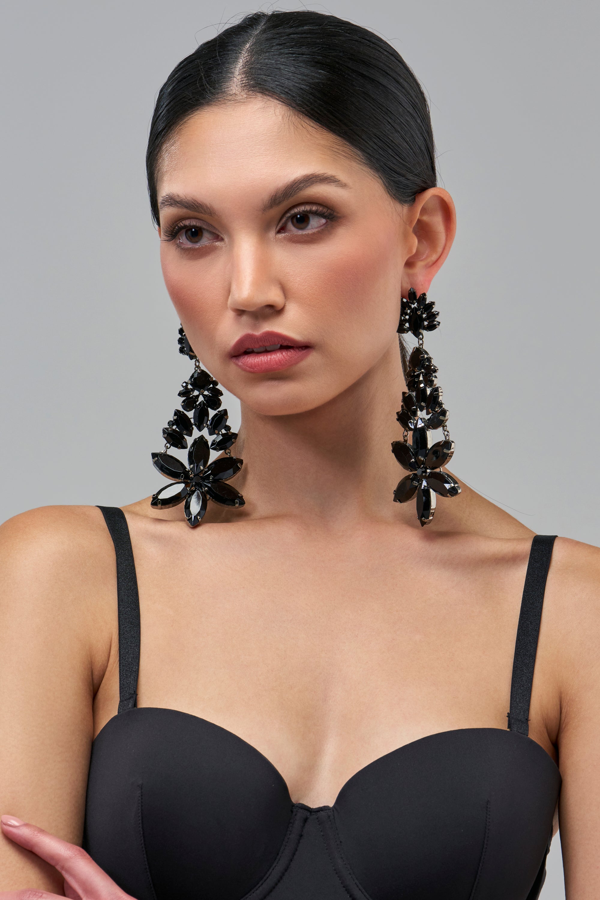 A model wears MIDNIGHT ORCHID CHANDELIER EARRINGS, which are long, black rhinestone earrings featuring tiered, flower-shaped accents.