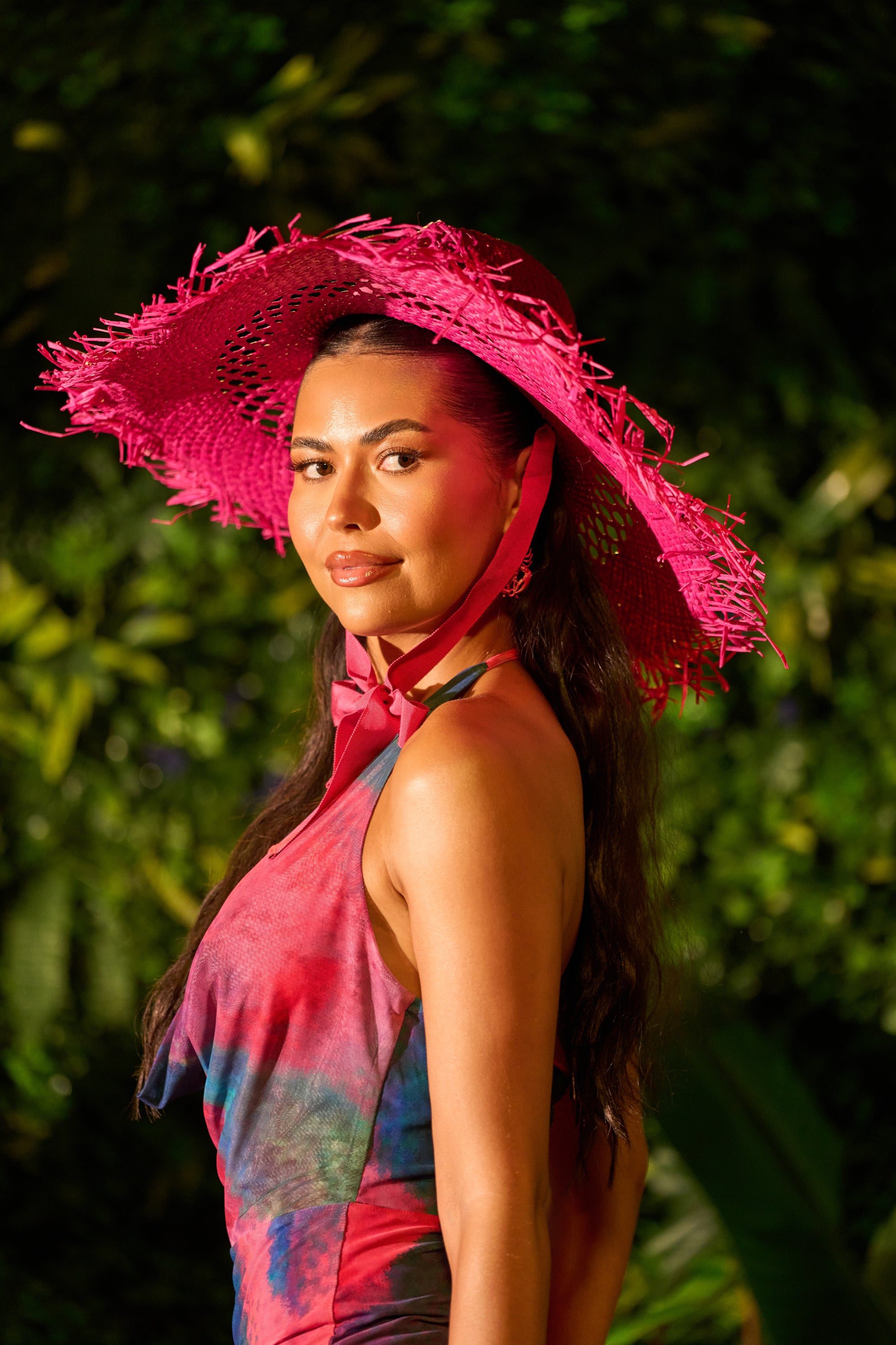 A woman poses confidently outdoors, smiling slightly in lush greenery, wearing the INTO THE SUN HAT— a bright pink straw hat—paired with a colorful tie-dye dress.
