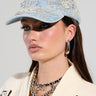 A person wearing a BLESSING IN DENIM EMBELLISHED BASEBALL HAT, adorned with pearls and intricate embroidery. Their long hair complements large hoop earrings, layered silver necklaces, and a cream-colored top with graphic patches.