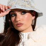 A woman is wearing the BLESSINGS ONLY EMBELLISHED BUCKET HAT in light blue denim, along with hoop earrings. Her long brown hair complements a white textured coat featuring a studded collar. She gazes to the side, gently touching the brim of her stylish hat with one hand.