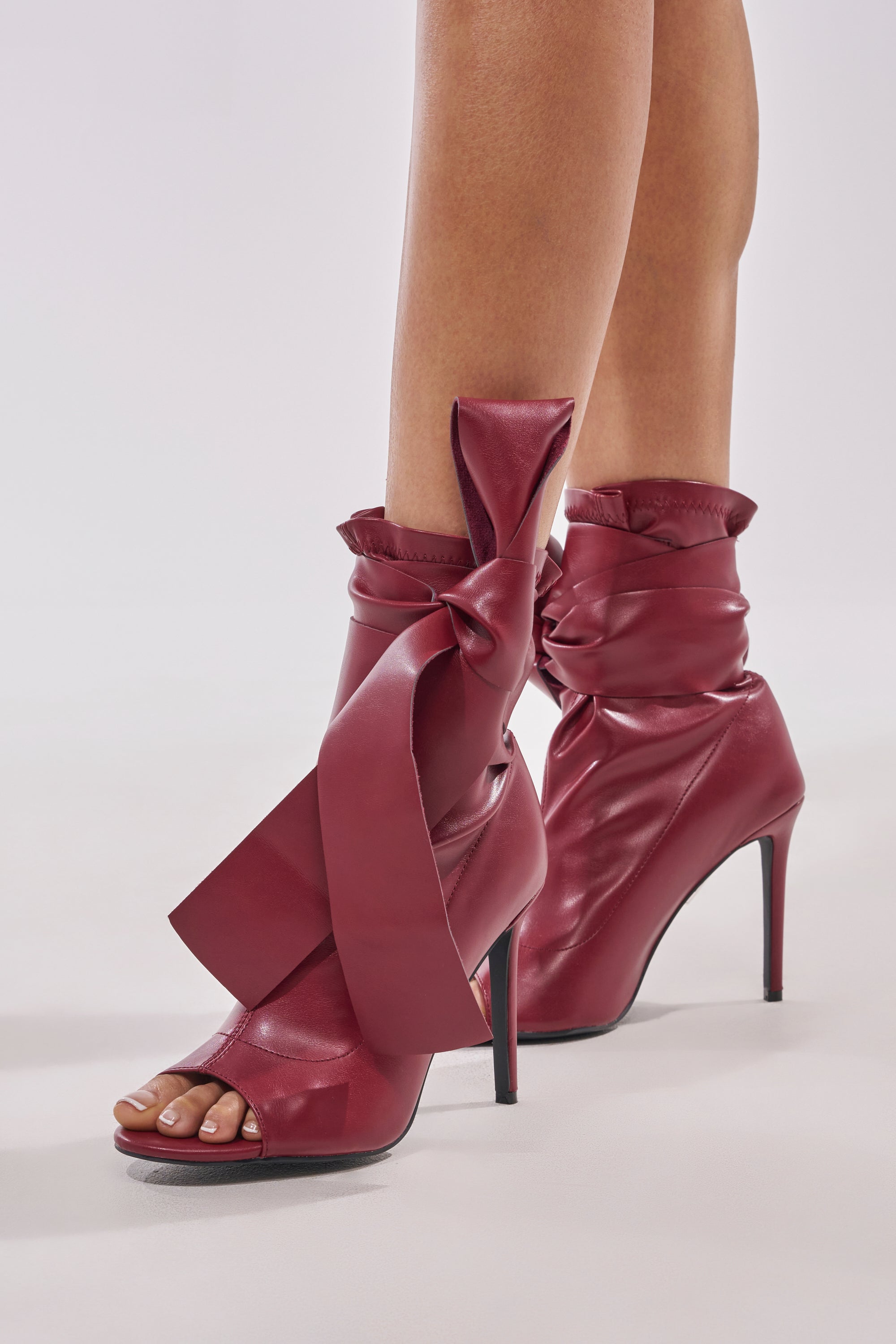 A close-up of a person wearing the AZALEA WANG KLARICE BURGUNDY STILETTO BOOTIE—high-heeled, open-toe boots in shiny vegan leather, featuring dramatic ankle bows, an almond peep toe, and elegant stiletto heels.