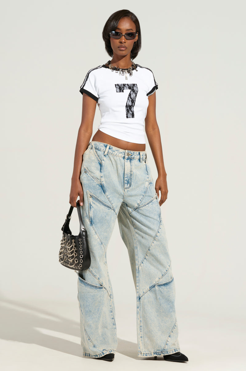 A person poses confidently in a white t-shirt with the number "7" and the SNAP OUT OF IT DENIM PANT, featuring a baggy, low-rise wide-leg design. They accessorize with a patterned handbag and sunglasses, set against a plain background that highlights their stylish outfit.