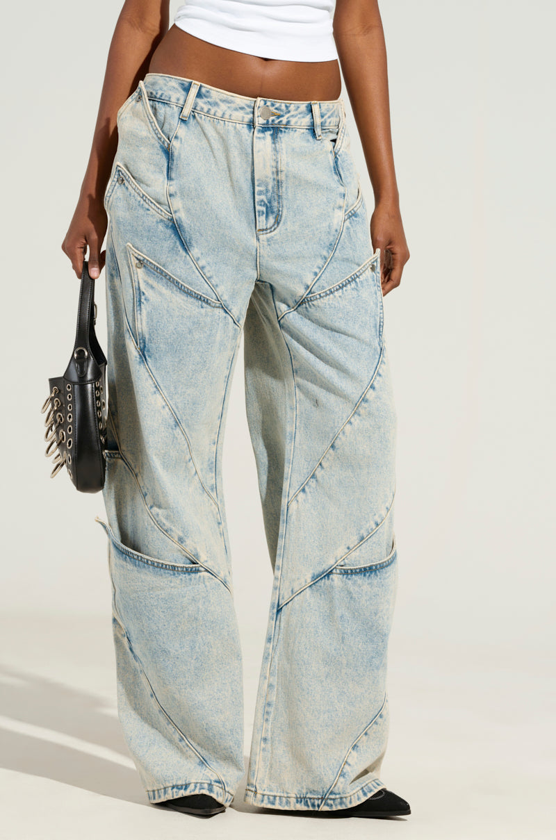 The image features a person from the waist down wearing the SNAP OUT OF IT DENIM PANT, which are low-rise wide-leg jeans in light blue with a white waistband. They are holding a black handbag embellished with silver spikes against a plain, light-colored background.