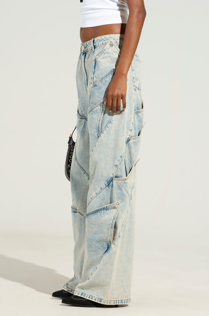 A person wearing the SNAP OUT OF IT DENIM PANT, featuring a low-rise wide-leg design with a pattern of overlapping sections, paired with a white crop top. The outfit is accented with stylish silver snap button fastenings and accessorized with a small black purse, creating an overall casual and chic look.