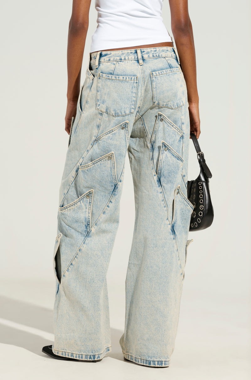 A person is styled in the SNAP OUT OF IT DENIM PANT, showcasing intricate zigzag stitching with its light-wash, low-rise wide-leg design. They pair it with a white top and carry a black bag embellished with silver grommets, creating a casual yet trendy look.
